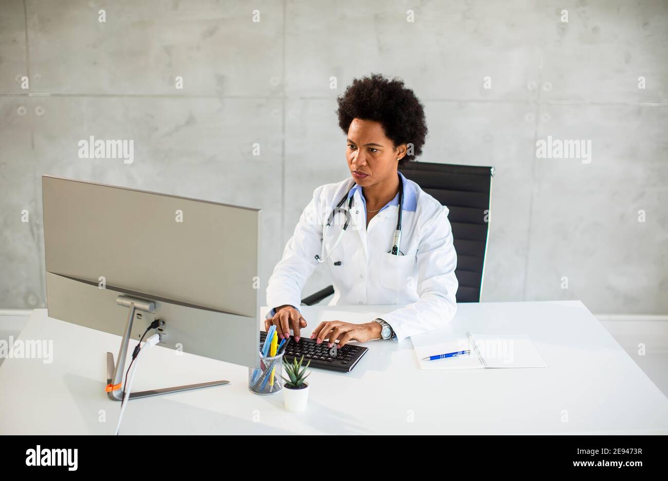 Femme afro-américaine médecin portant un manteau blanc avec stéthoscope assis derrière le bureau Banque D'Images