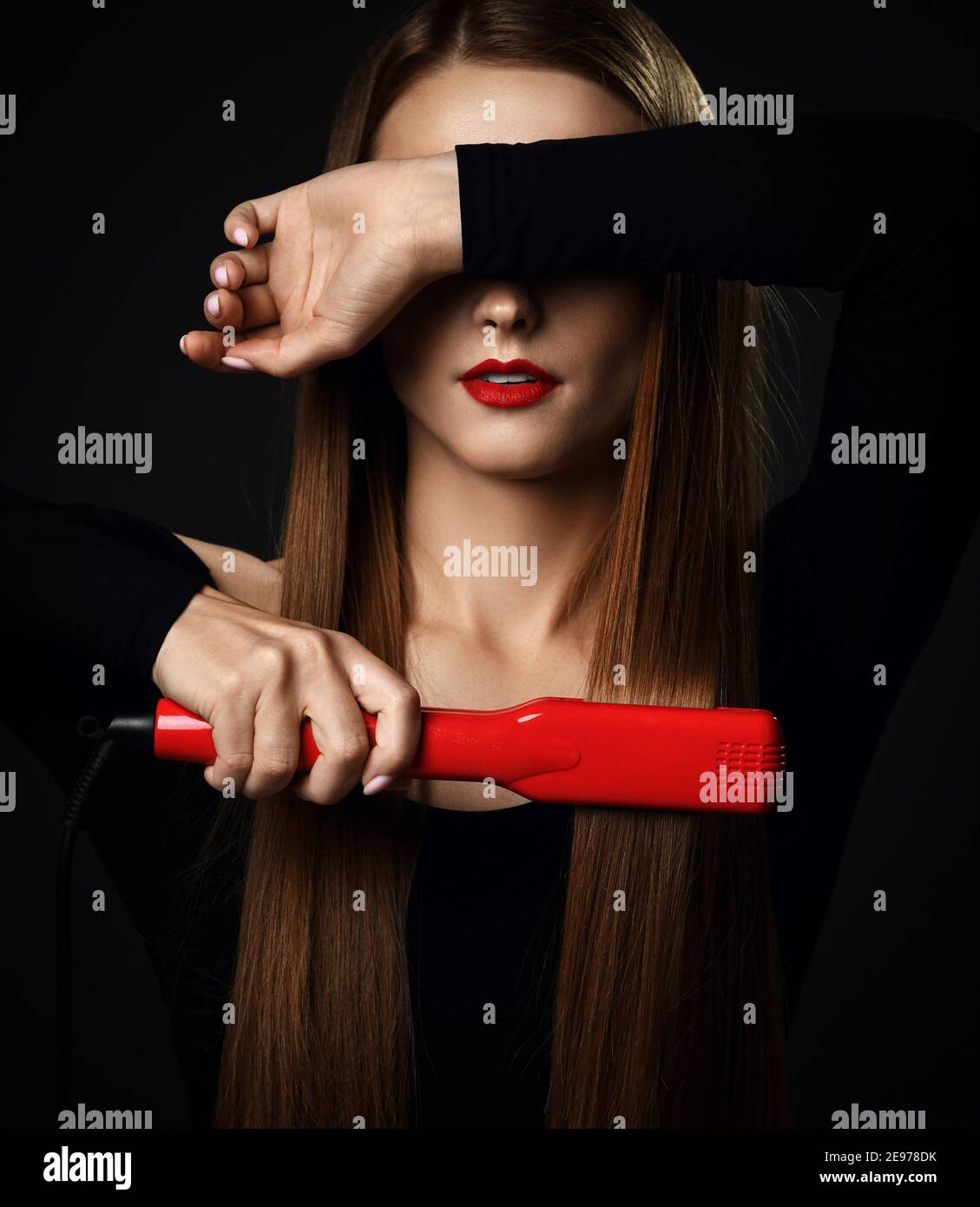 Jeune jolie femme avec de longs cheveux soyeux dans des vêtements noirs utilise un lisseur rouge et couvre les yeux de l'autre main Banque D'Images