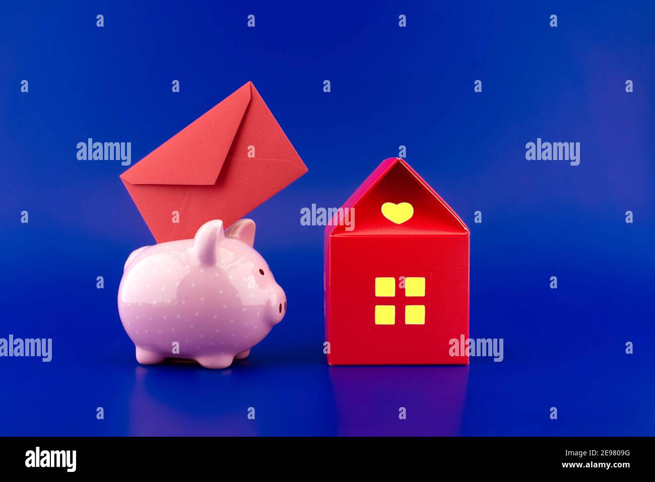 Enveloppe rouge festive insérée dans une tirelire rose à côté d'une petite maison rouge de papier avec chaud lumière dans les fenêtres sur fond bleu avec cop Banque D'Images