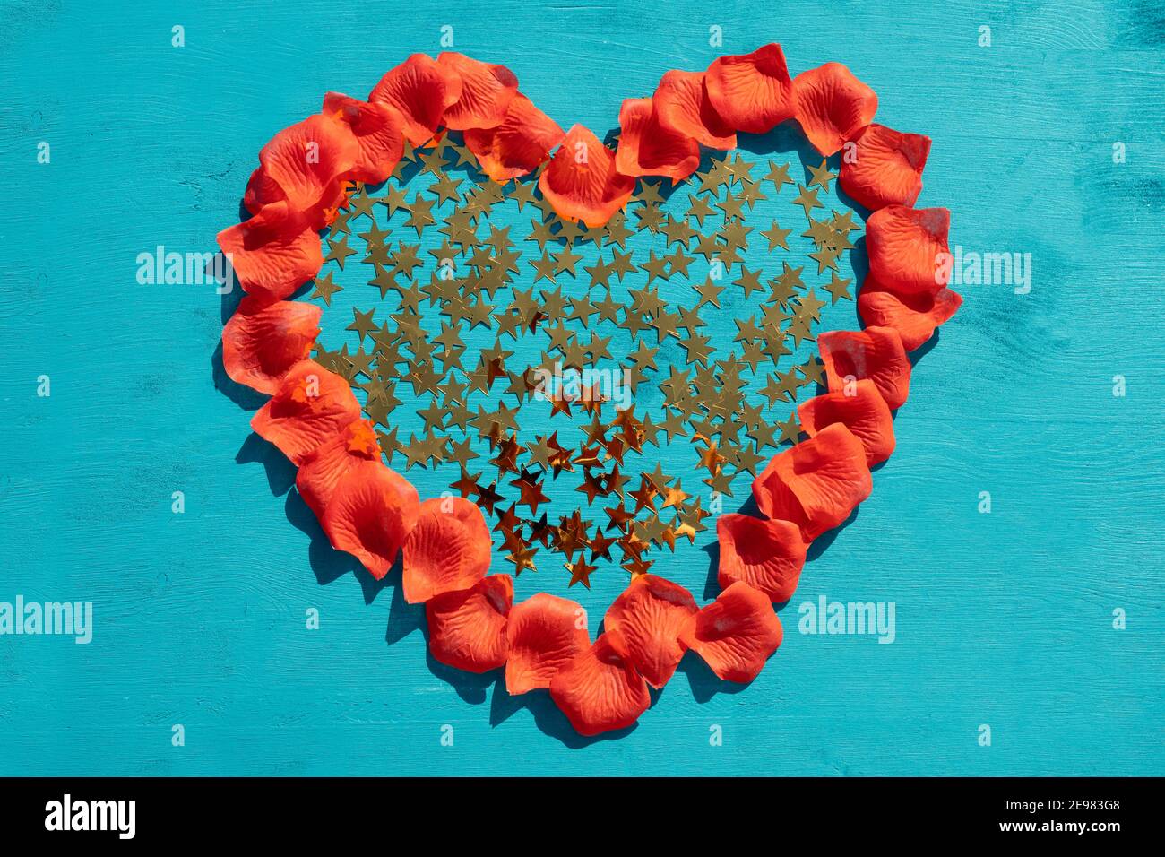 Love and Valentines Day concept : vue d'en haut sur un coeur en forme de feuilles roses rouges. Le coeur est rempli d'étoiles métalliques d'or sur bois bleu Banque D'Images