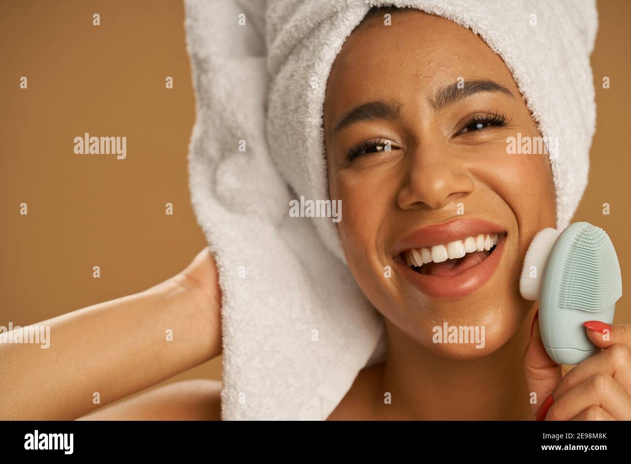 Gros plan de la jeune femme joyeuse après la douche regardant excitée de l'appareil photo tout en utilisant la brosse de nettoyage du visage en silicone, posant isolé sur fond beige. Beauté, concept de soins spa Banque D'Images