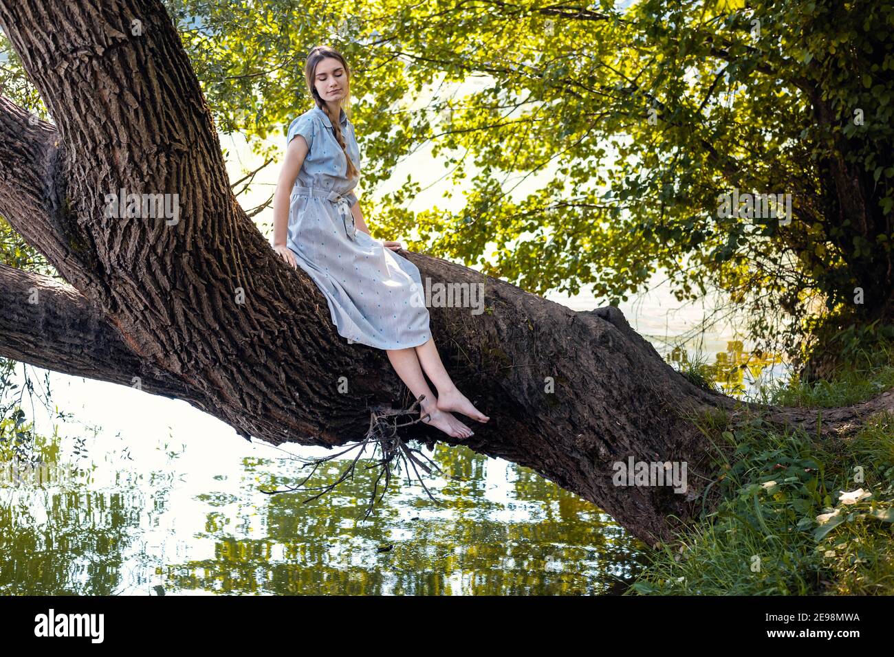 Jeune fille en robe d'été bleue assise sur un arbre au-dessus de la rivière Banque D'Images