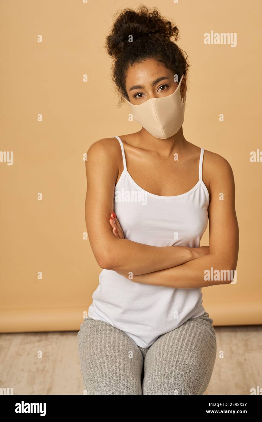 Photo en studio d'une jeune femme de race mixte vêtue de vêtements décontractés portant un masque de protection pour le visage en regardant l'appareil photo tout en posant les bras croisés sur un fond beige. Sécurité, concept de pandémie Banque D'Images