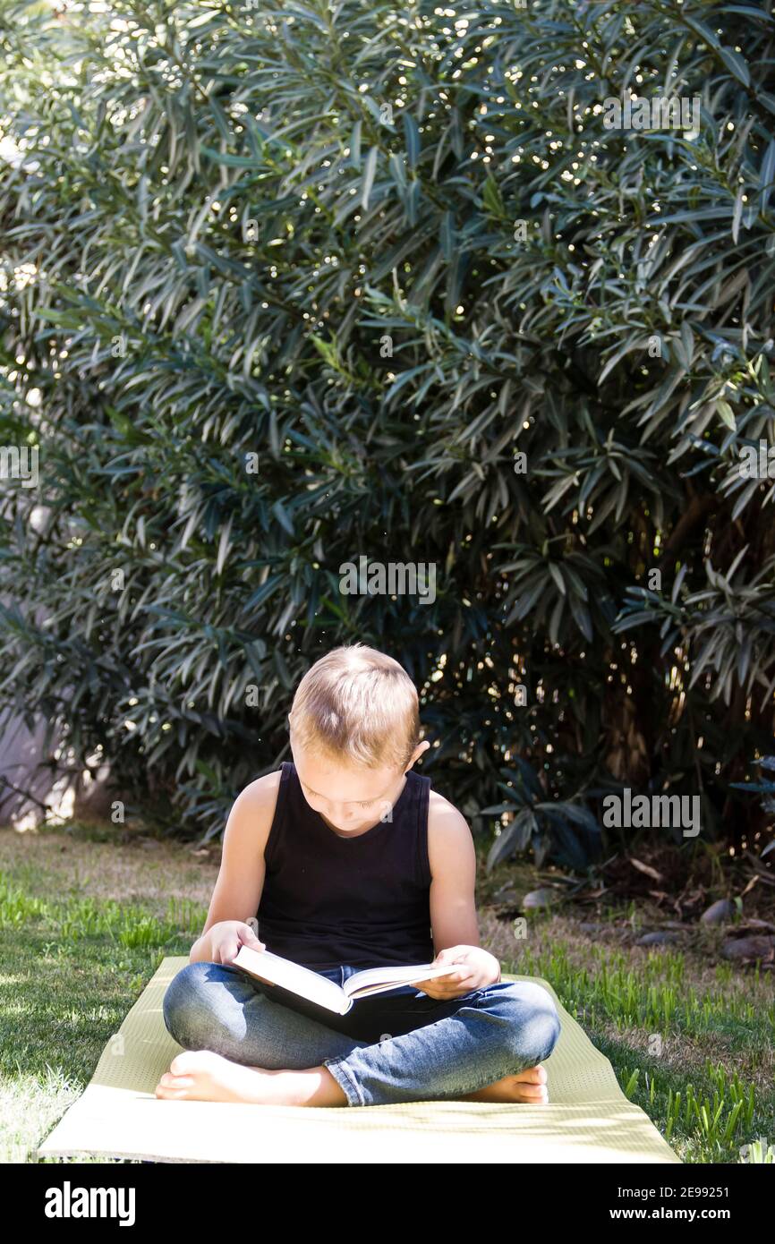 Le petit garçon est assis avec le livre sur le tapis de yoga dans le jardin. Banque D'Images