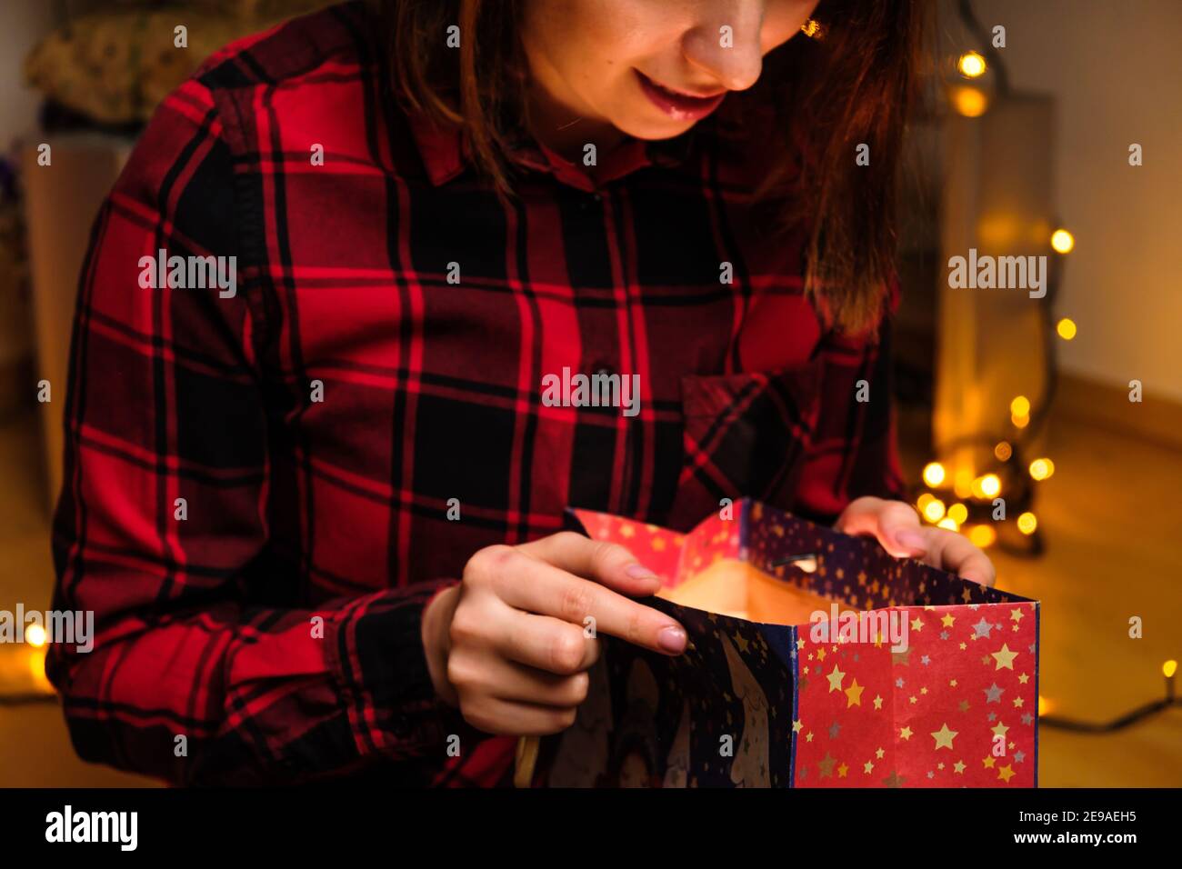 Une femme heureuse dans une chemise rouge ouvre un cadeau de vacances dans le paquet avec la lumière à l'intérieur. Concept anniversaire ou fête de la Saint-Valentin. Banque D'Images