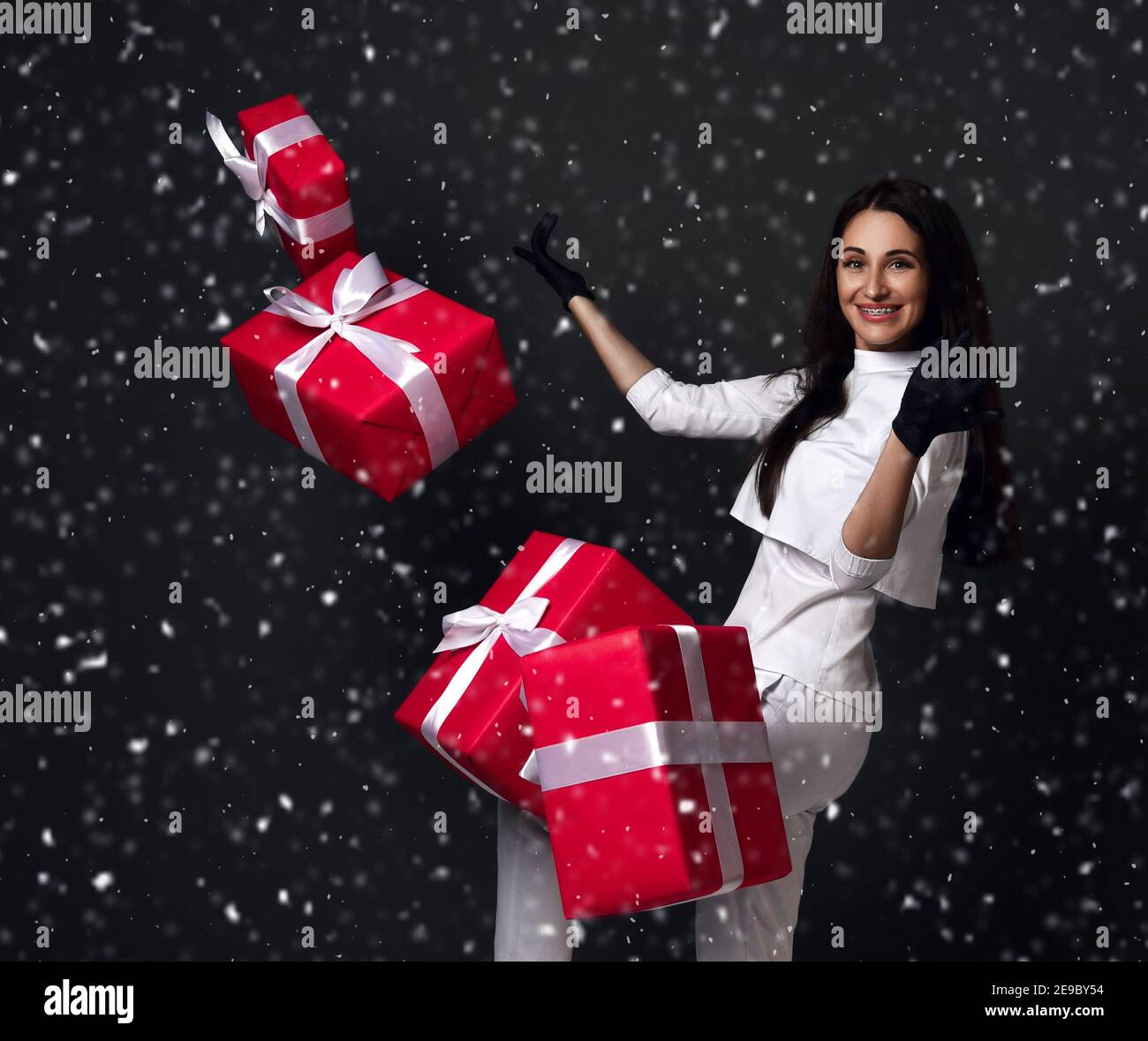 Jeune femme souriante médecin dentiste en uniforme blanc et en latex gants avec bretelles debout et boîtes-cadeaux volantes autour Banque D'Images