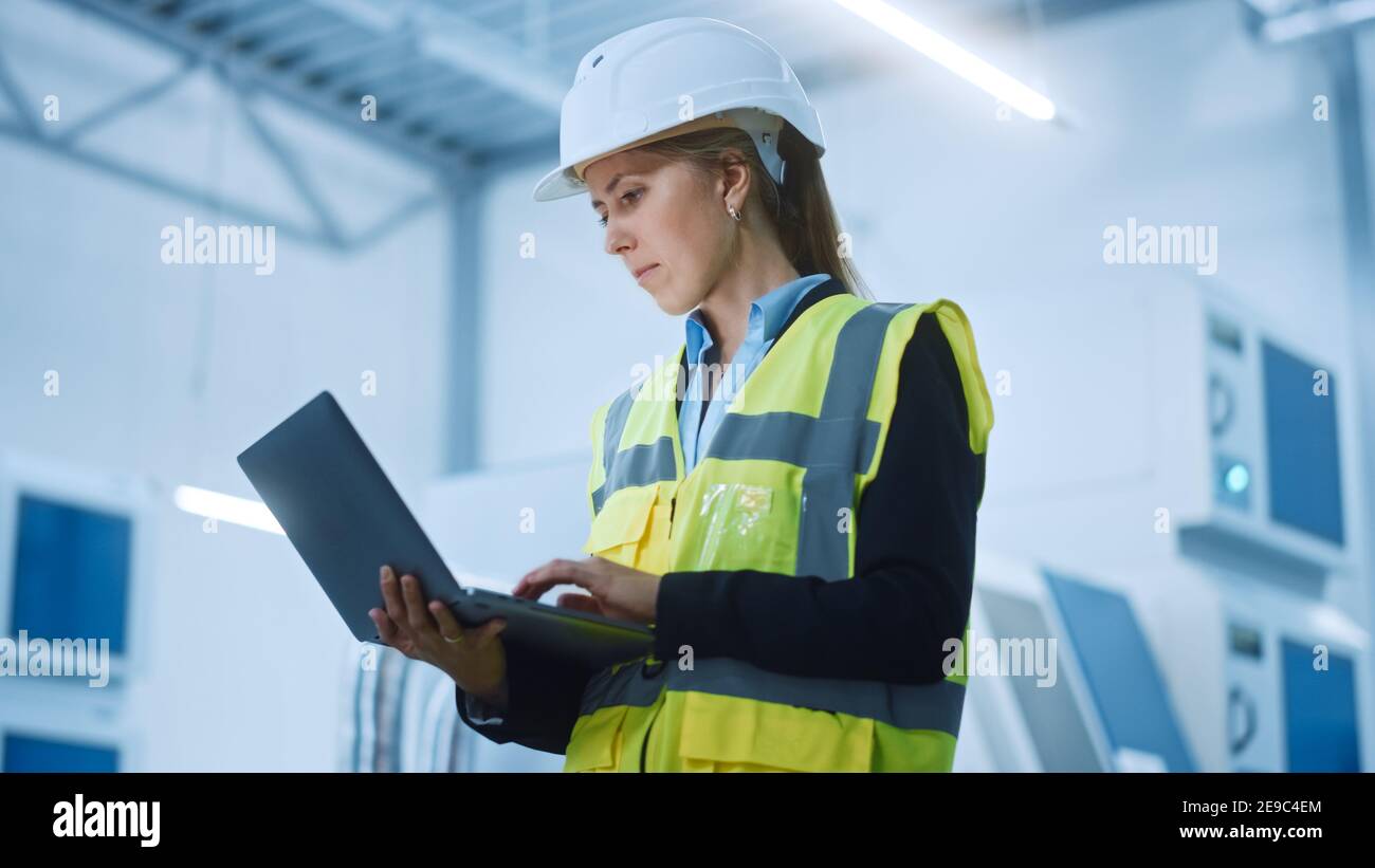 Usine de haute technologie : femme ingénieure confiante et professionnelle portant une veste de sécurité et un casque de sécurité tenant et travaillant sur un ordinateur portable. Moderne et lumineux Banque D'Images
