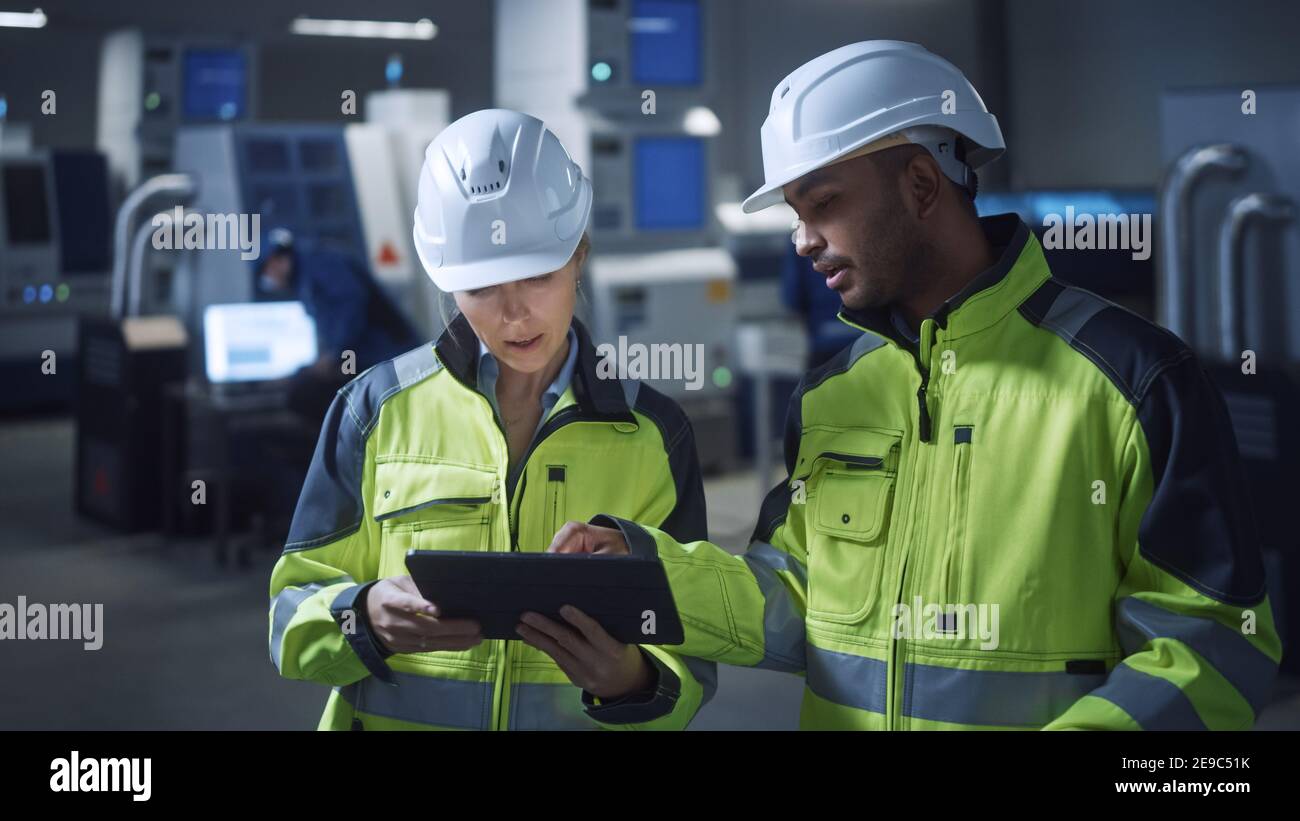 Ingénieur en chef et chef de projet portant des gilets de sécurité et des chapeaux durs, utiliser une tablette numérique dans l'usine moderne, parler, optimiser la production Banque D'Images