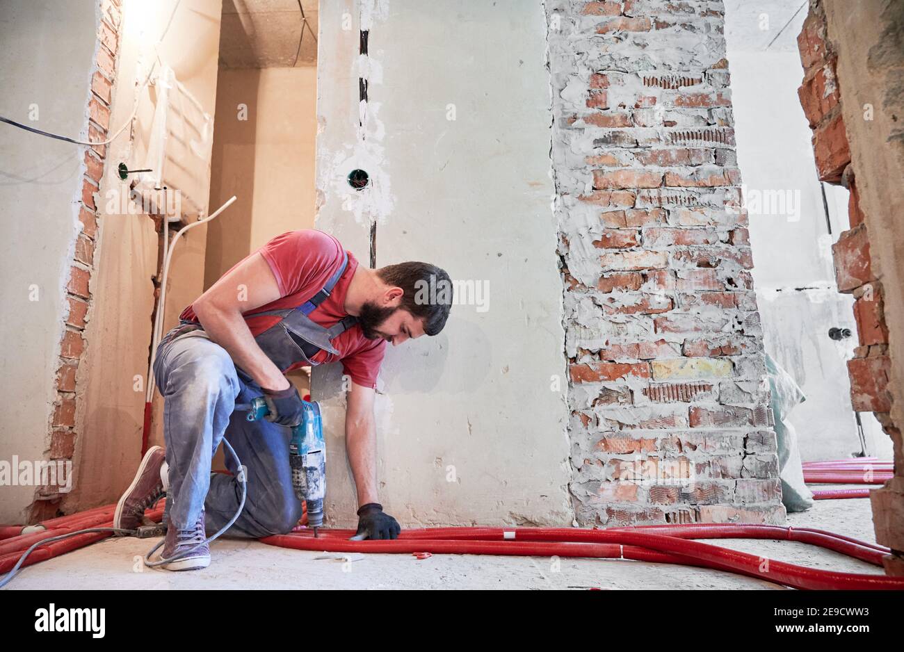 Vue horizontale du plombier barbu installant un système de chauffage au sol dans une pièce vide avec des murs en briques. Homme portant une combinaison et des gants, debout sur un genou, travaillant avec perforateur Banque D'Images