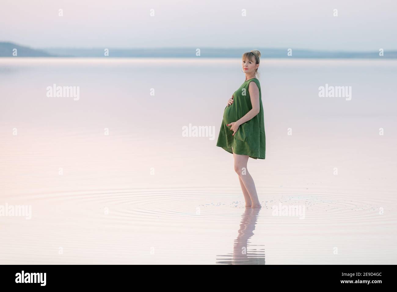 Femme enceinte élégante dans la robe verte posant au coucher du soleil, regardant l'appareil photo. Coucher de soleil incroyable sur un lac peu profond. Banque D'Images