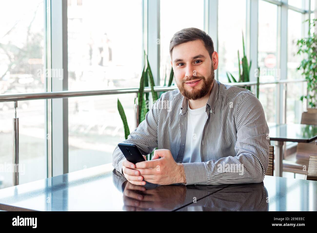 Souriant homme caucasien envoyant un message sur smartphone dans le café seul près de la fenêtre Banque D'Images