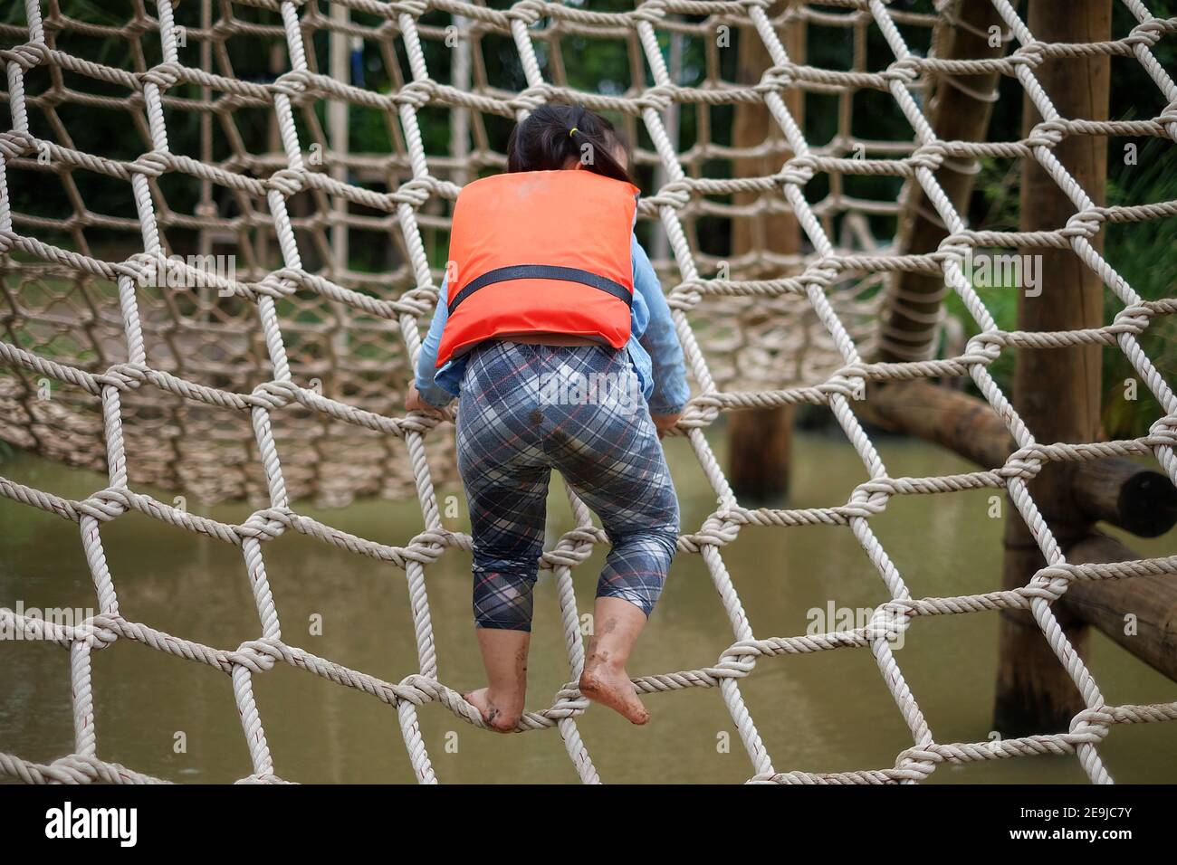 Une jeune fille asiatique mignonne portant un gilet de sauvetage orange monte un filet d'escalade dans un parcours d'obstacles dans un camp d'été. Banque D'Images