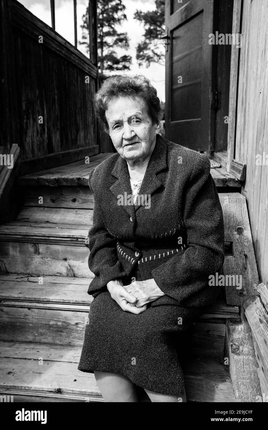 Une vieille femme s'assoit près d'une maison rurale. Photo en noir et blanc. Banque D'Images