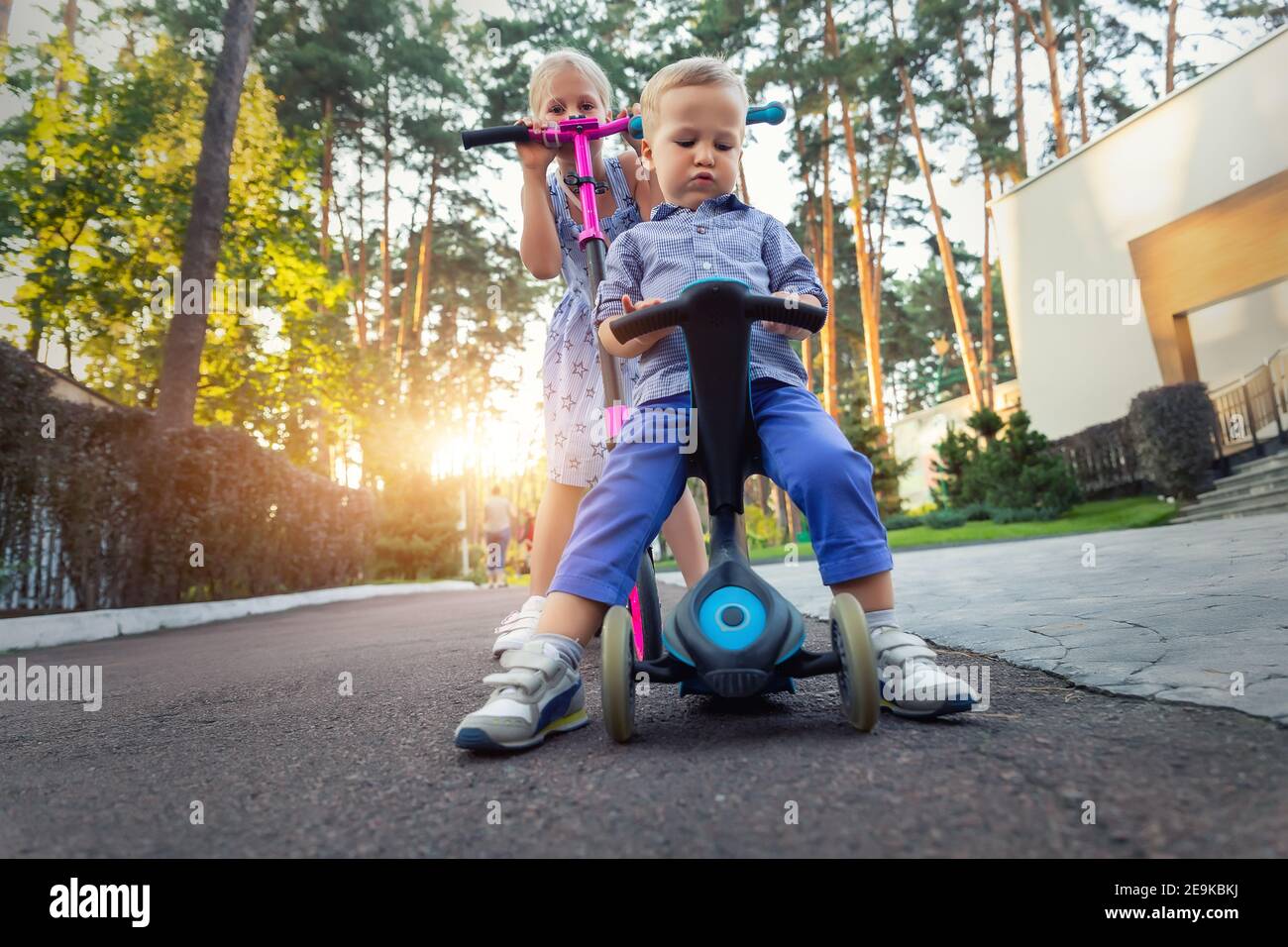 Deux adorables petits frères et sœurs caucasiens garçons en chemise et fille en robe ayant plaisir à monter trois roues équilibre vélo de course scooter dans la forêt du parc de la ville Banque D'Images