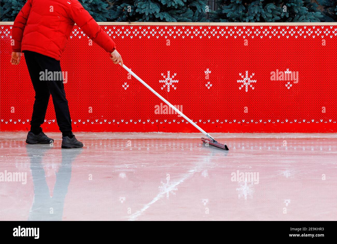 Un ouvrier de stade de glace, entouré d'une clôture rouge et portant une veste rouge, frottant la surface de glace avec une vadrouille caoutchoutée pour enlever les petits éclats. Copier Banque D'Images