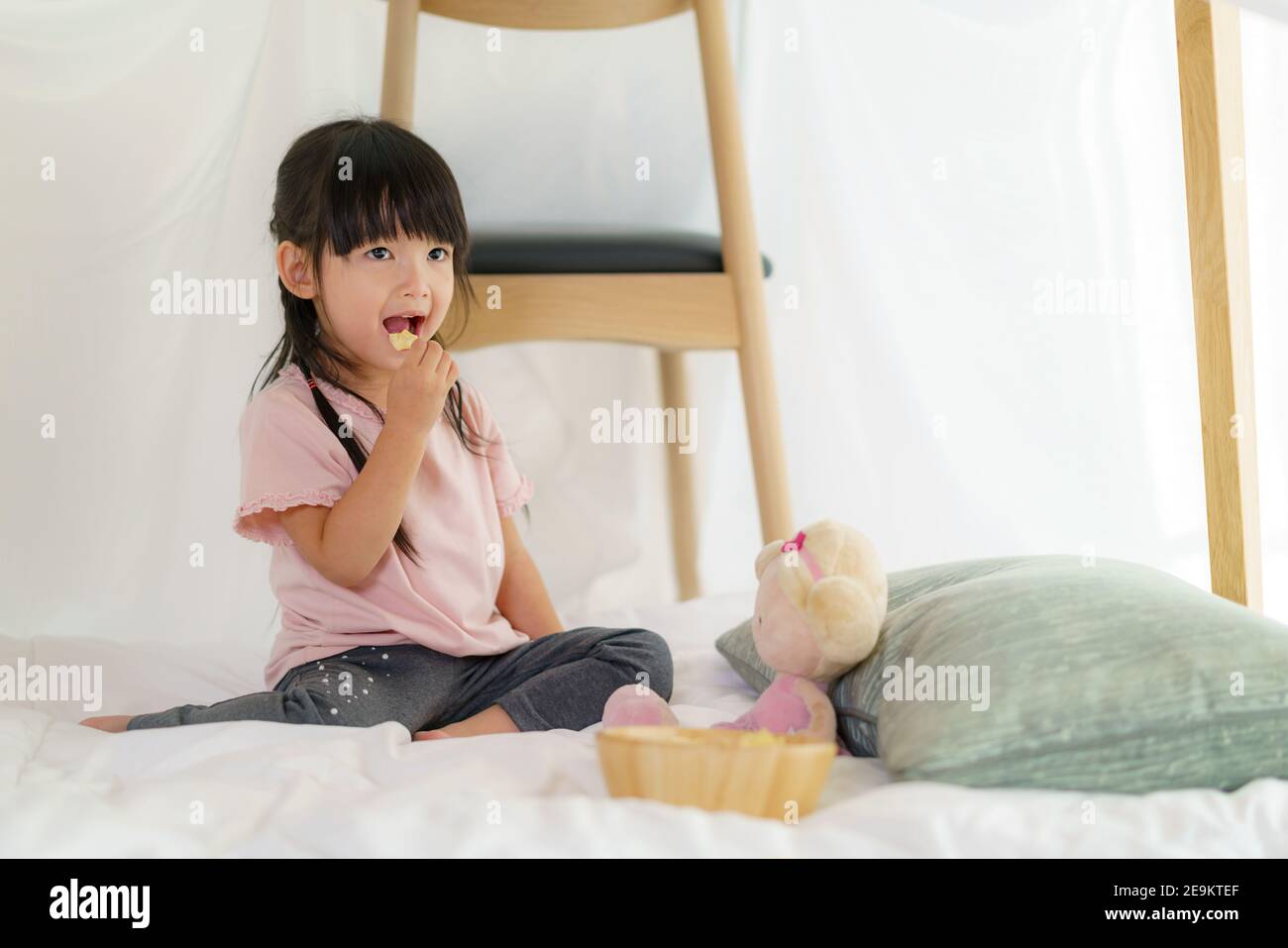 Asiatique mignon petite fille manger snack tout en s'asseyant dans un couverture fort dans la salle de séjour à la maison pour un refuge parfait loin de leurs autres membres de la famille un Banque D'Images