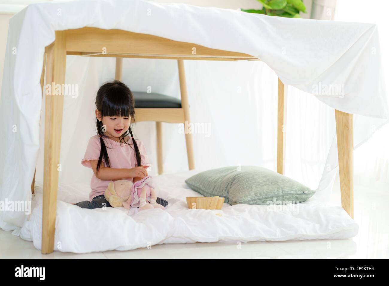 Asiatique mignon petite fille jouer sa poupée tout en s'asseyant dans une couverture de fort dans le salon à la maison pour parfait éloignez-vous des autres membres de leur famille a Banque D'Images
