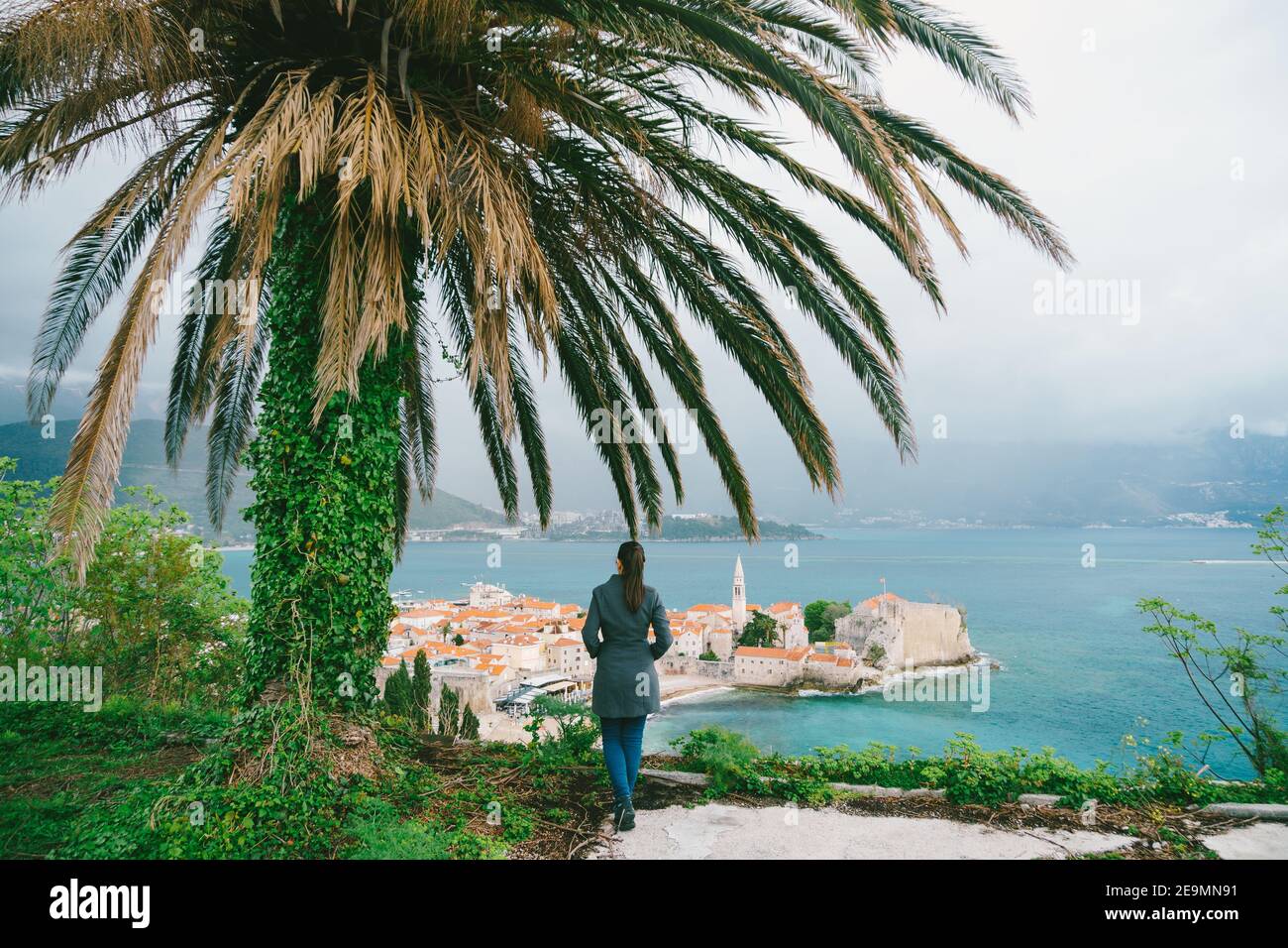 La femme est debout sous un palmier avec son dos À la caméra regardant la vieille ville de Budva Au Monténégro Banque D'Images