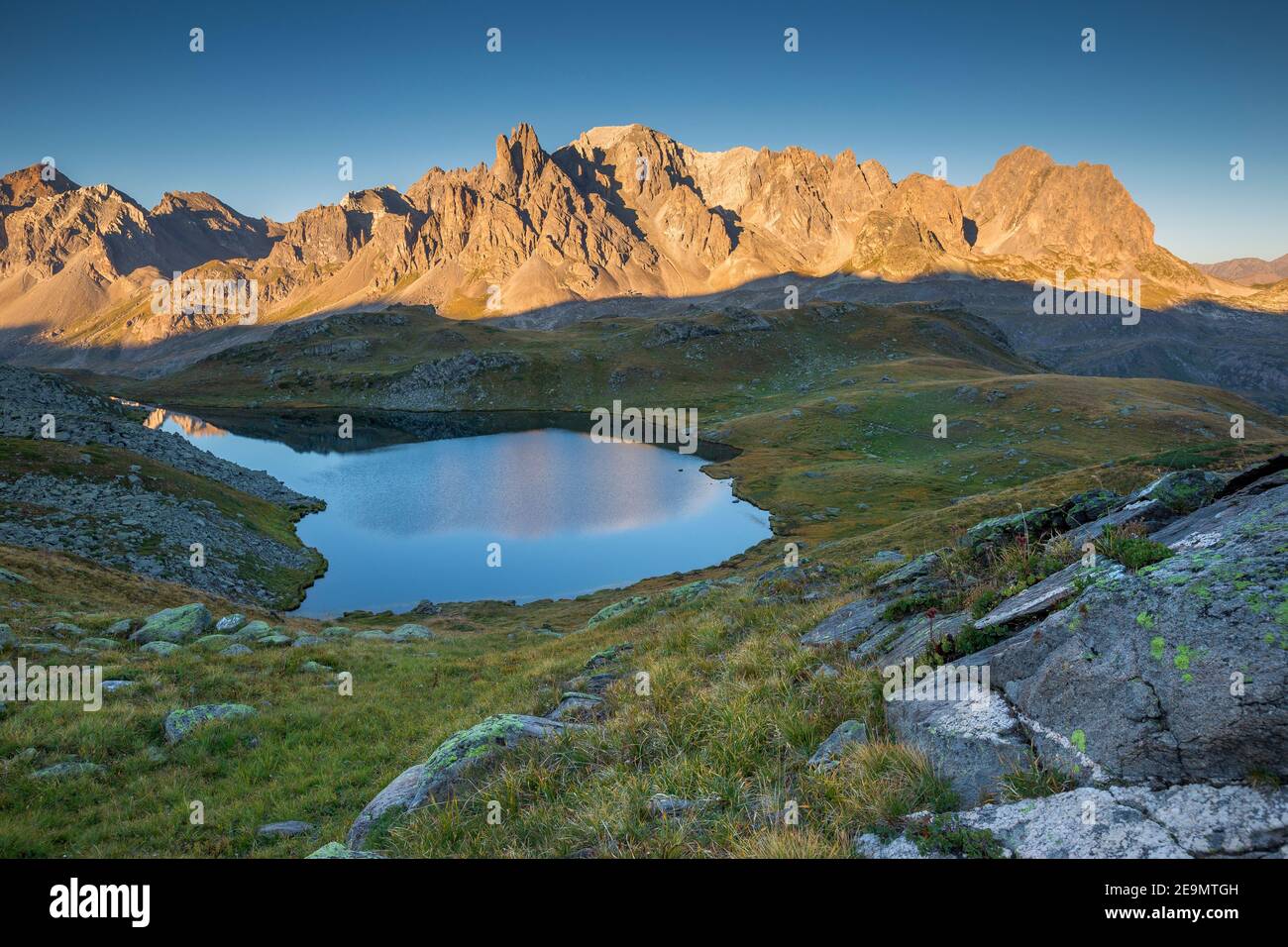 Vue panoramique sur les prairies verdoyantes, lac alpin bleu (Lac long), pics de montagne au soleil levant. Vallée de la Clarée. Névache, Hautes Alpes. Banque D'Images