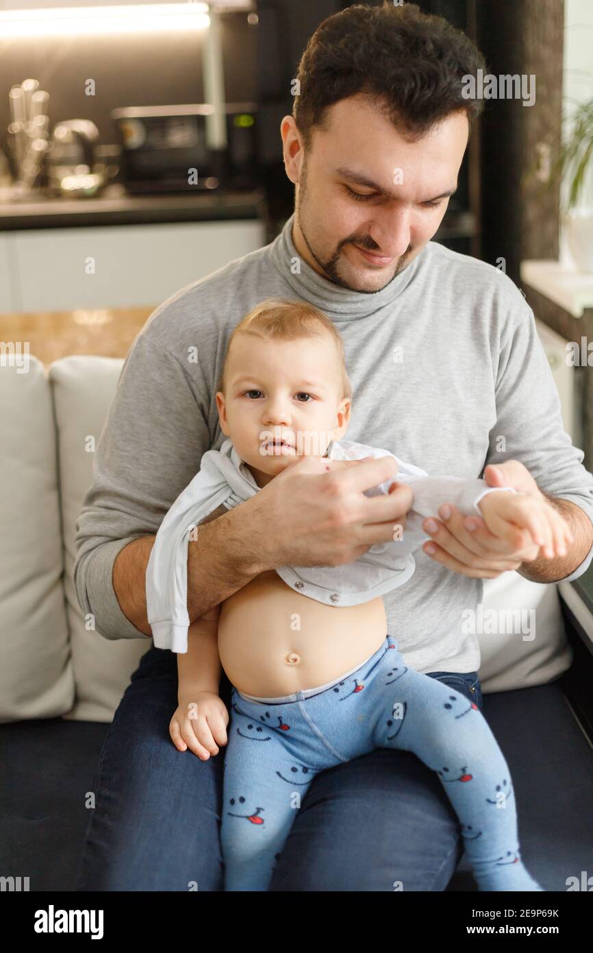 Le père habille le bébé dans la salle de séjour, assis sur un canapé à la  maison. Vêtements sous-chemises et curseurs de bébé sur bébé gros plan.  Concept de soins à domicile