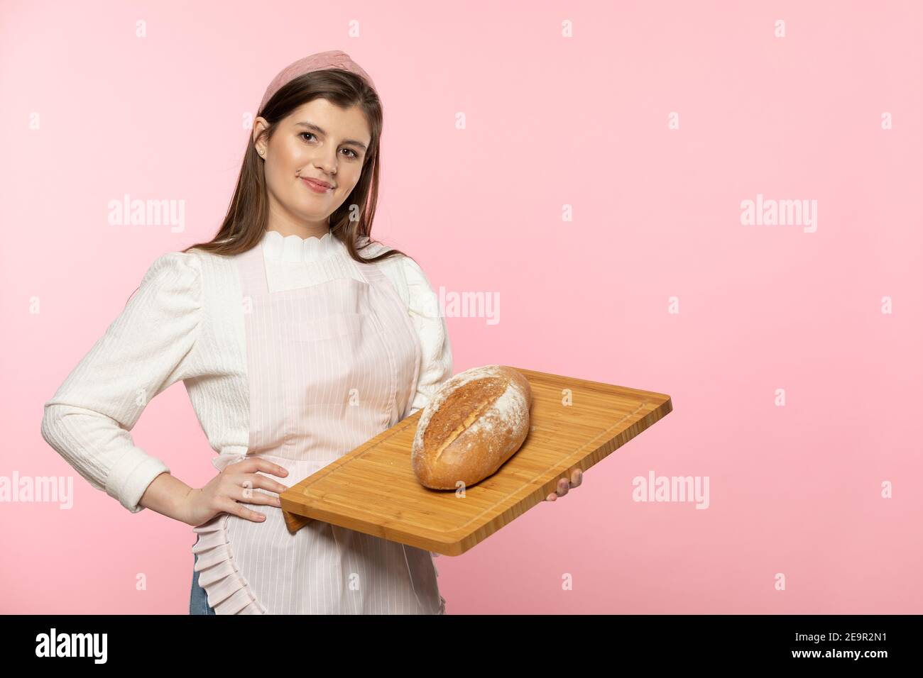 Une jeune fille de belle beauté européenne tient dans ses mains un pain fraîchement cuit sur une planche de bois. Toute la composition est fixée sur un fond rose clair uniforme. Banque D'Images