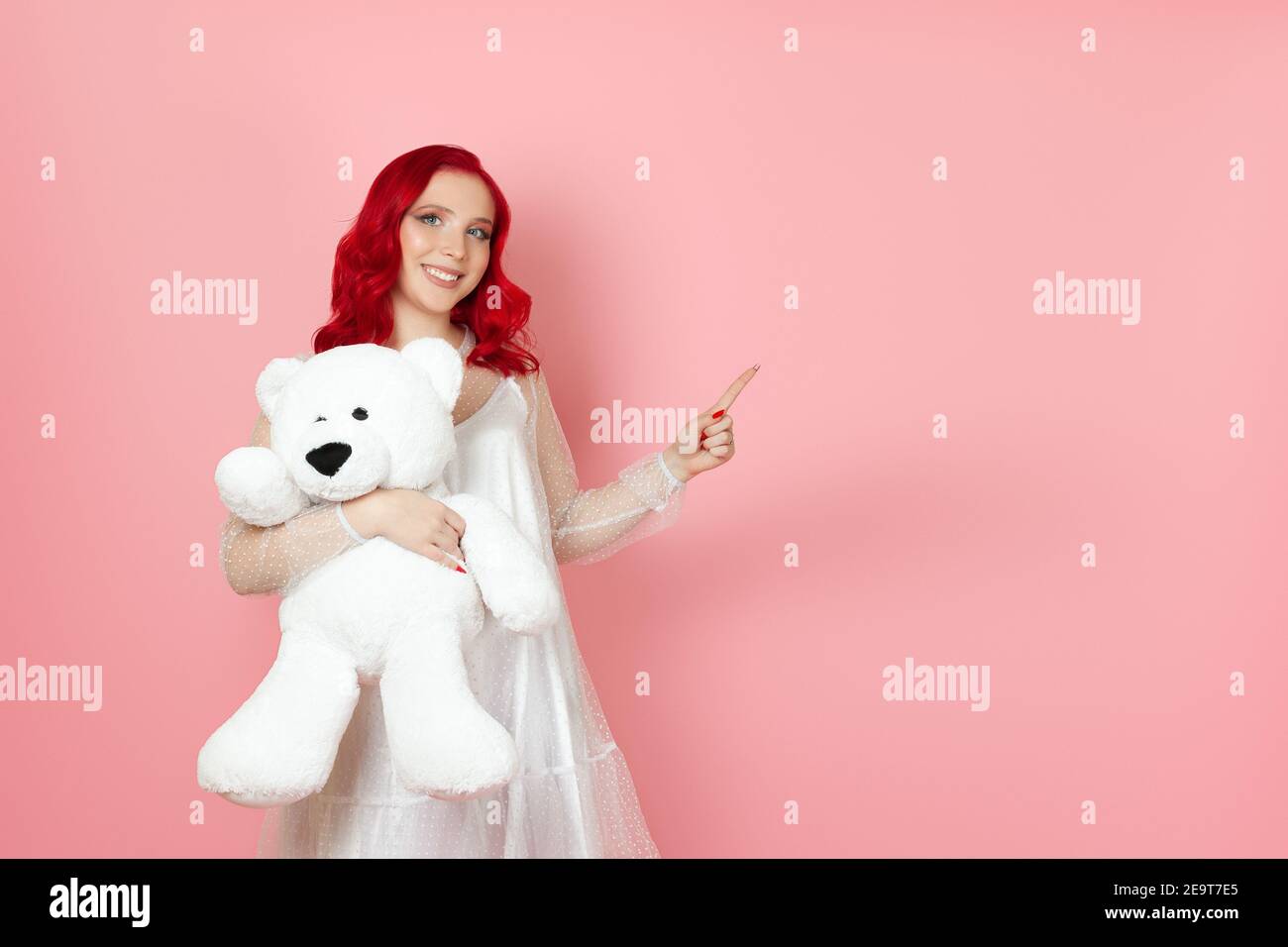 femme avec les cheveux rouges tient grand ours blanc teddy et pointe avec son index vers un espace vide pour le texte Banque D'Images