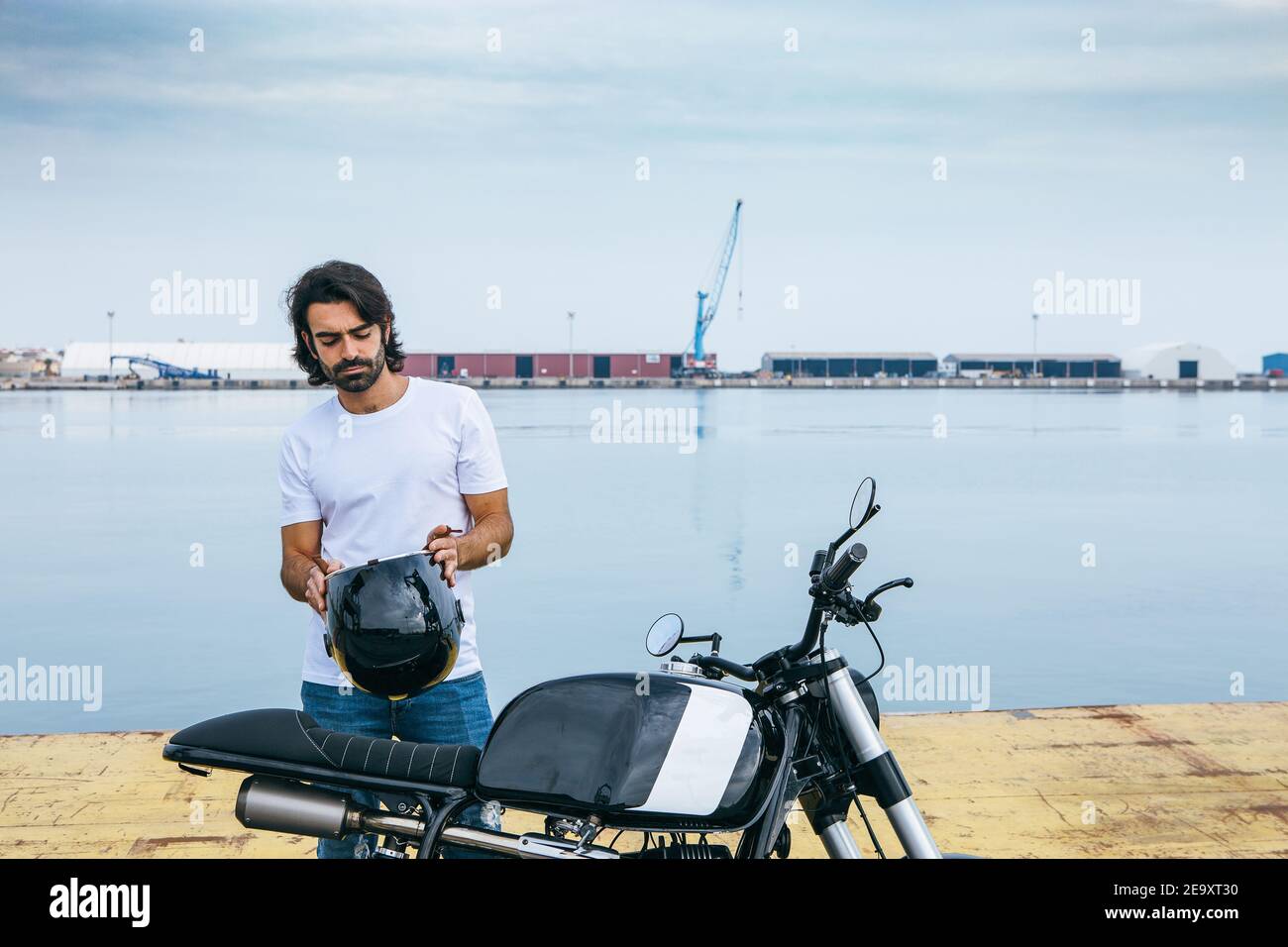 Jeune homme ethnique à barbe confiant en t-shirt blanc et jeans en moto debout au bord de la mer Banque D'Images