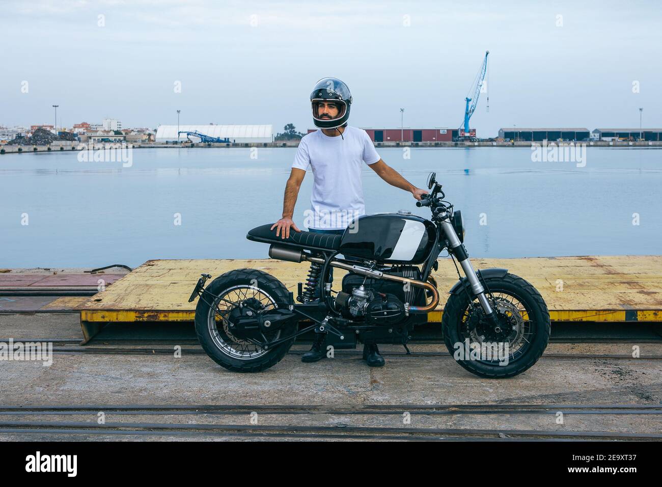 Jeune homme ethnique à barbe confiant en t-shirt blanc et jeans en moto debout au bord de la mer Banque D'Images