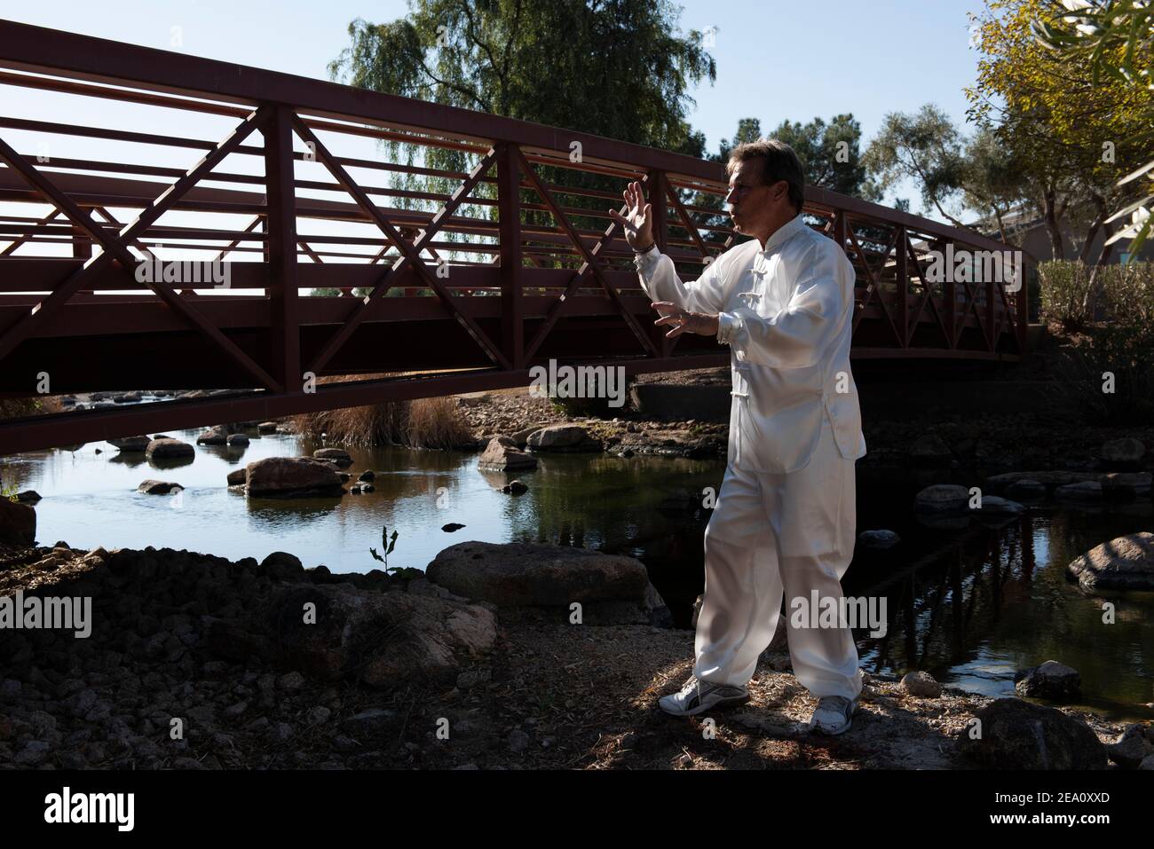 Homme portant un costume blanc et exécutant le Tai Chi sur un rocher dans un cadre extérieur avec un pont en métal derrière lui et l'eau en arrière-plan. Banque D'Images