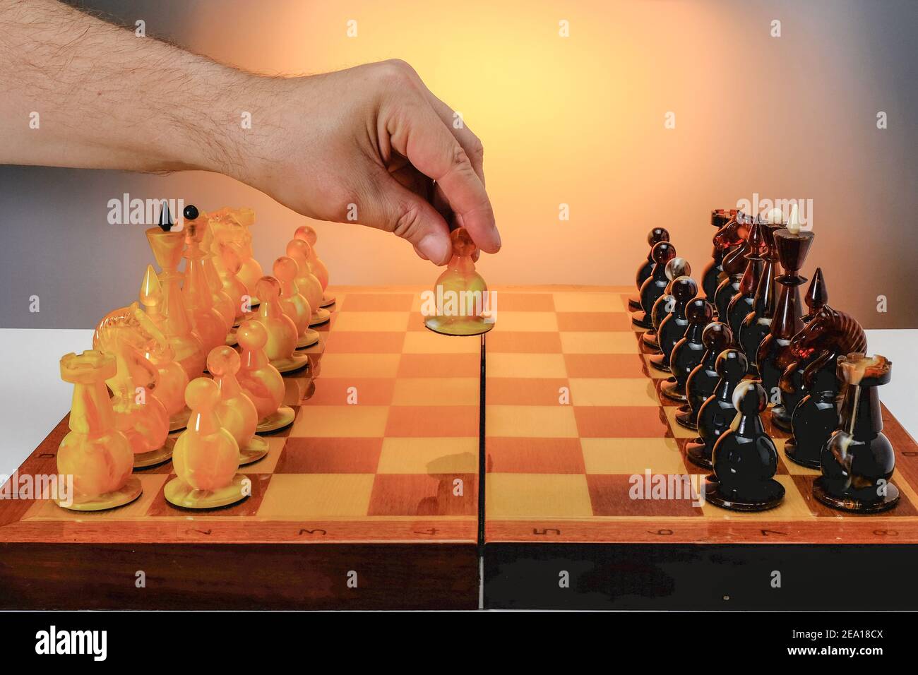 Jeu d'échecs avec des figures d'échecs ambres sur le plateau, premier mouvement. Main du joueur tenant une figure blanche. Avec fond doré dégradé. Banque D'Images