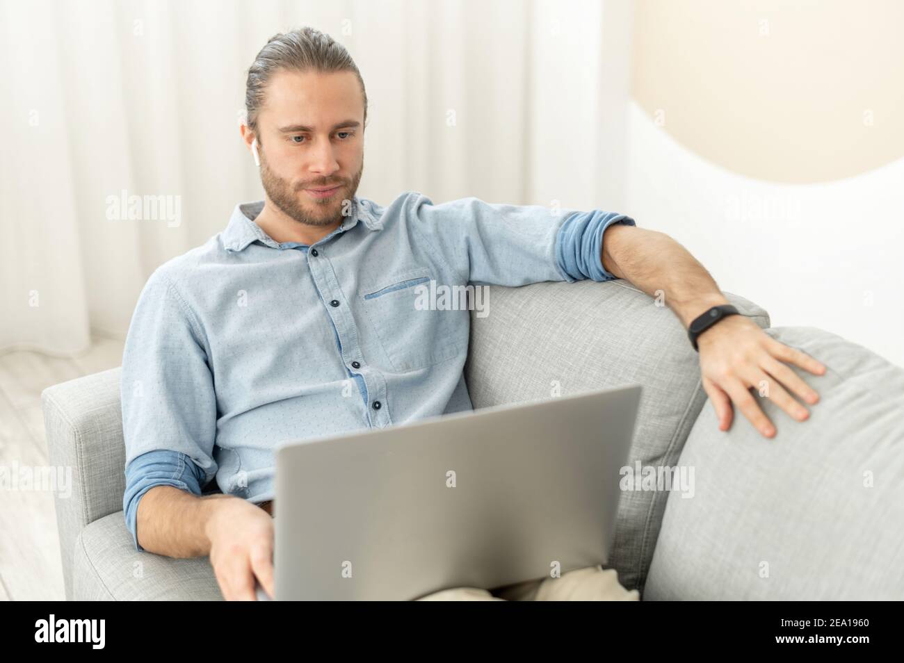 Un beau gars passe du temps dans les réseaux, un jeune homme allongé sur le canapé confortable et utilisant un ordinateur portable, regardant des films, travaillant à distance de la maison Banque D'Images