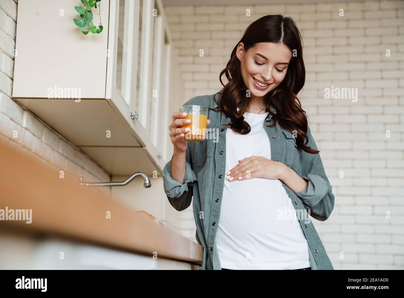 Bonne femme enceinte charmante souriante et buvant du jus dans confortable cuisine Banque D'Images