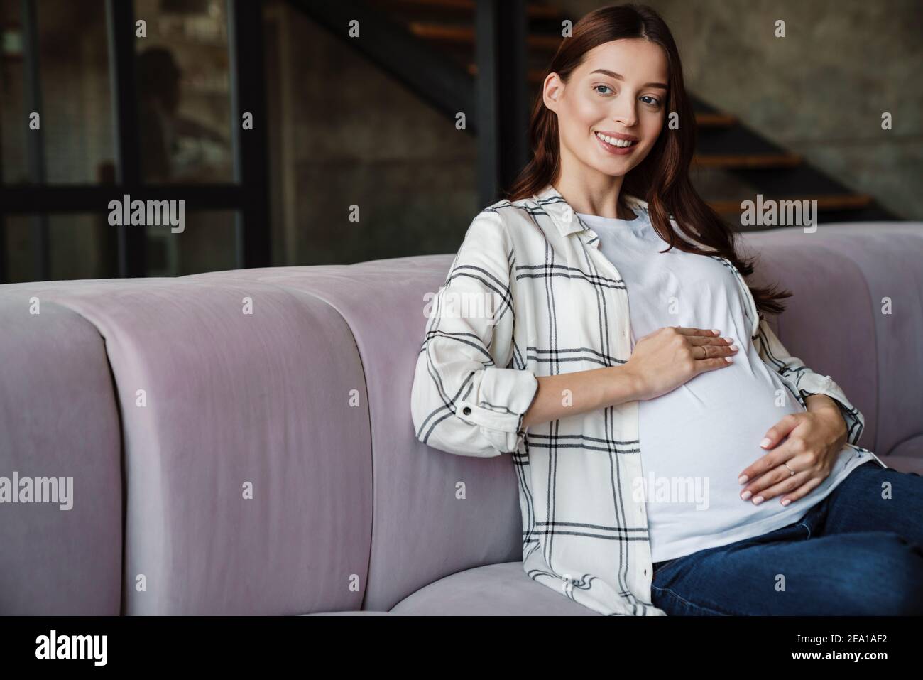 Bonne charmante femme enceinte souriante et assise sur un canapé chambre confortable Banque D'Images