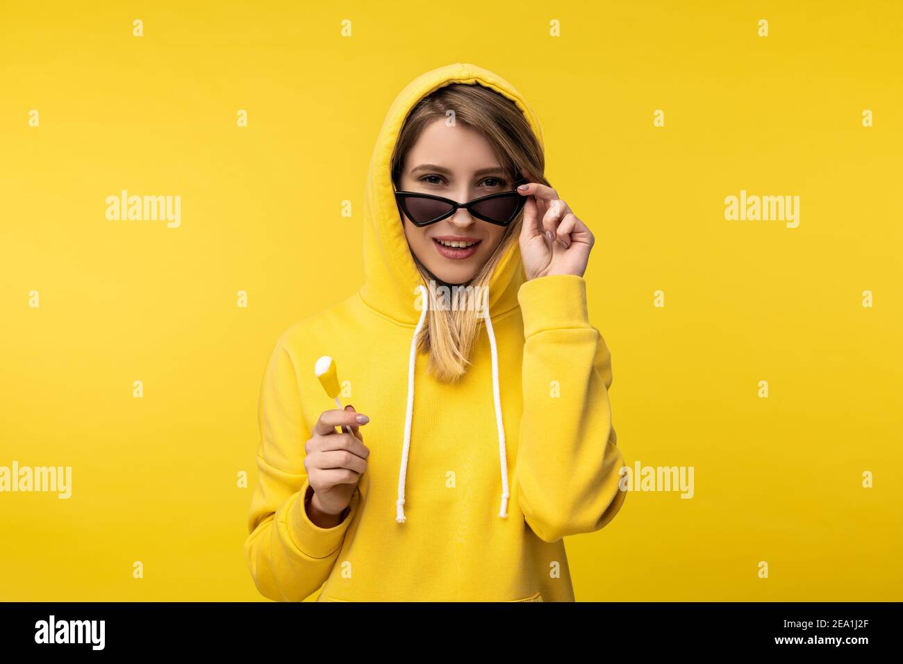 Photo de la jolie dame avec le Lollipop tient ses lunettes et le sourire. Porte un chandail jaune décontracté, un fond jaune isolé Banque D'Images