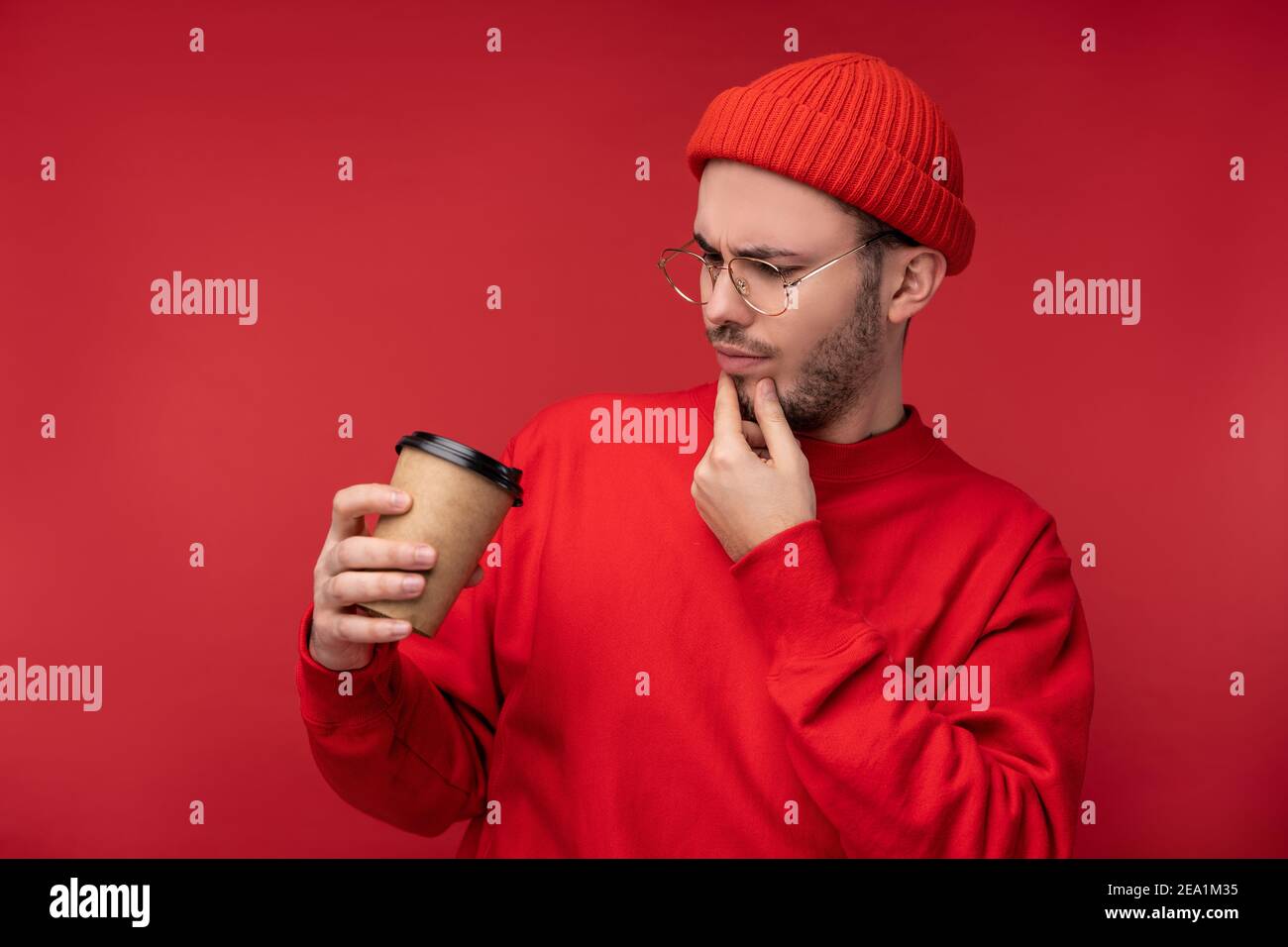 Photo d'un homme attrayant avec une barbe en lunettes et des vêtements rouges. Curieux homme regarde sa tasse de café, isolé sur fond rouge Banque D'Images