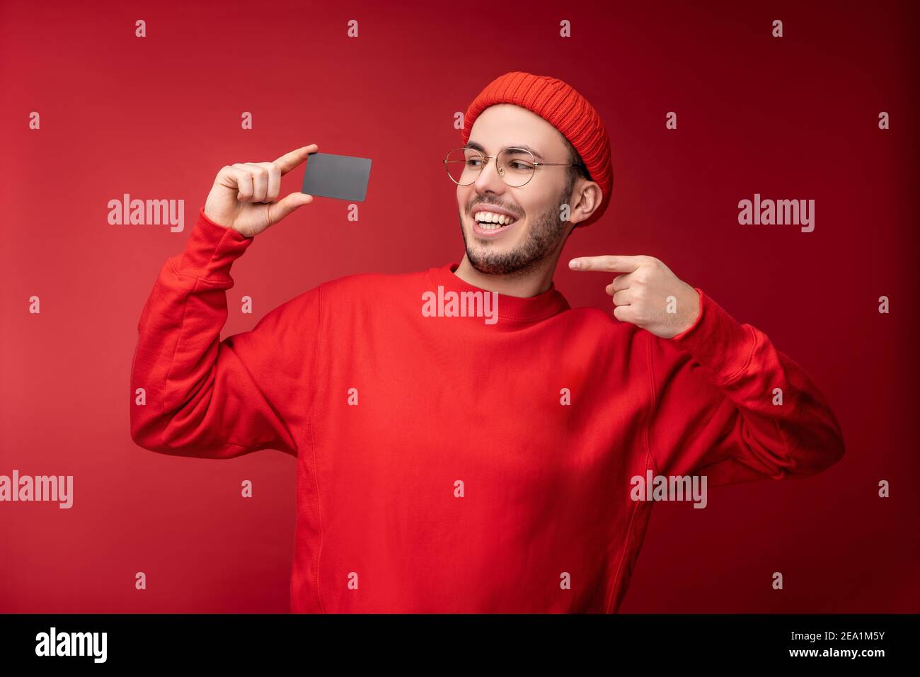 Photo d'un homme attrayant avec une barbe en lunettes et des vêtements rouges. Homme heureux tient et points à la carte de crédit, isolé sur fond rouge Banque D'Images