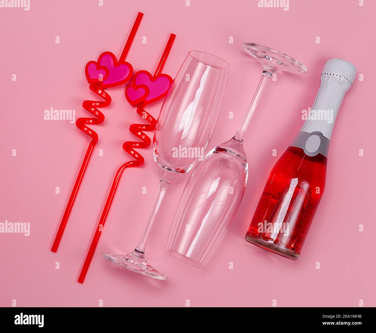 Deux verres à champagne avec pailles à boire en plastique rouge avec coeurs et bouteille de champagne rouge sur fond rose. Banque D'Images