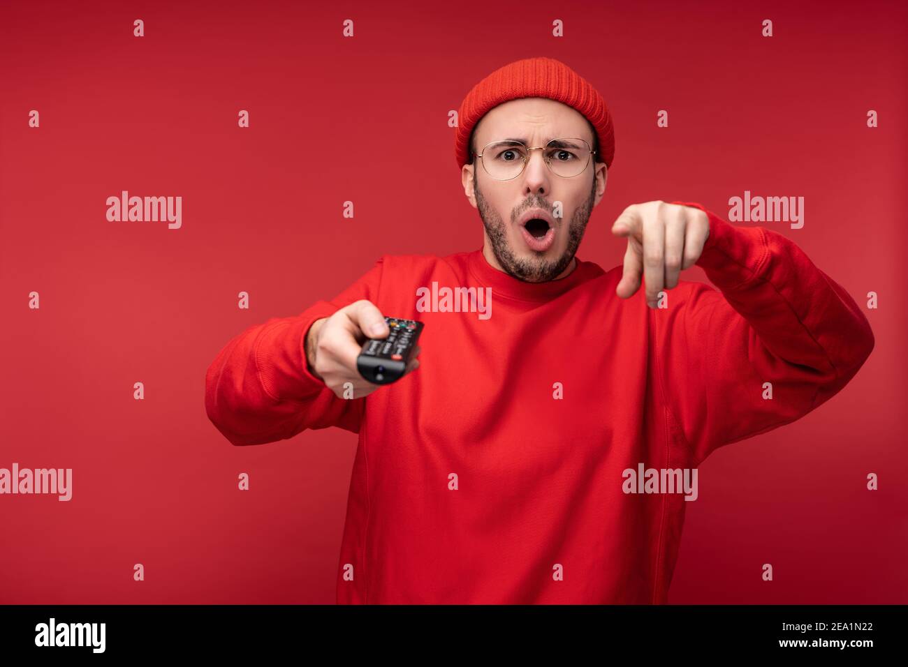 Photo de l'homme agité avec la barbe dans des vêtements rouges pointant les doigts vers l'appareil photo tient la console de télévision avec surprise et la bouche ouverte, isolée sur le rouge Banque D'Images