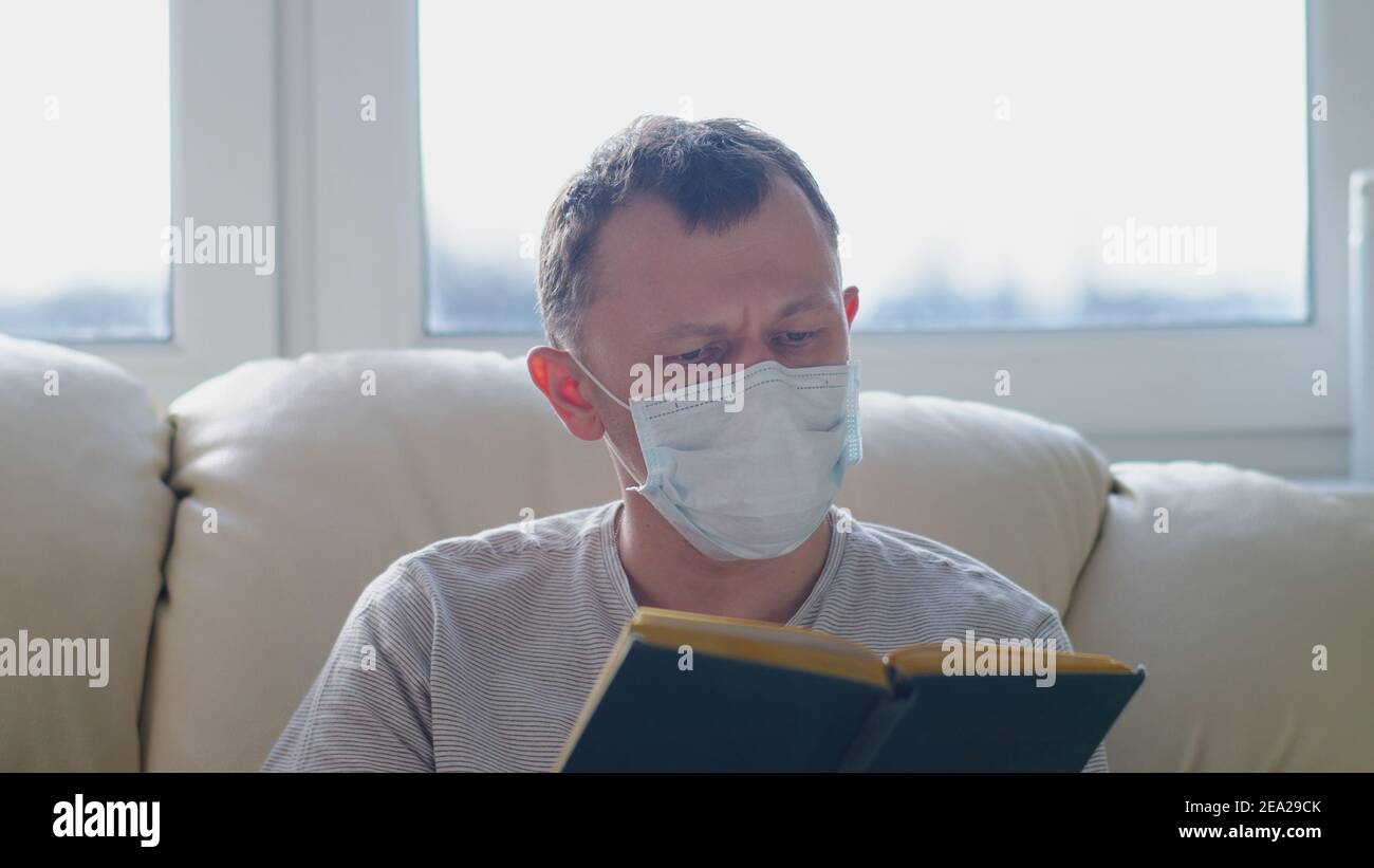 Homme attrayant dans le masque médical avec un livre dans son les mains regarde la caméra tout en étant assis sur un canapé Banque D'Images