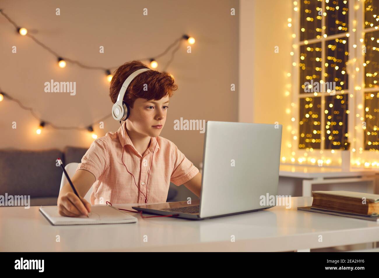 Un garçon aux cheveux rouges assis dans un casque, regardant une leçon ou un cours en ligne sur un ordinateur portable et prenant des notes Banque D'Images