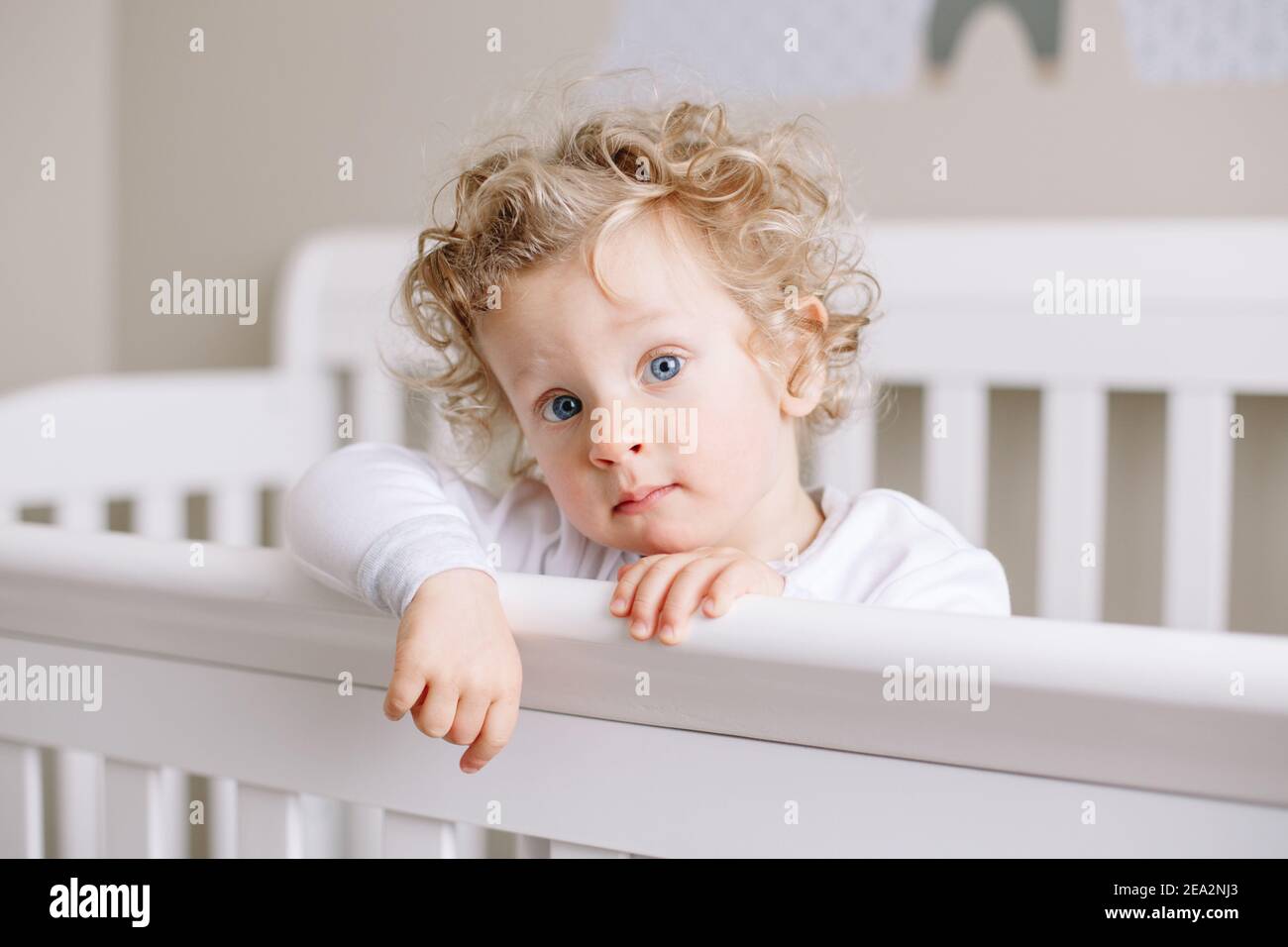 Mignon adorable bébé garçon tout-petit debout dans un berceau à la chambre des enfants à la maison. Curieux charmant petit garçon avec des cheveux blond et des yeux bleus regardant Banque D'Images