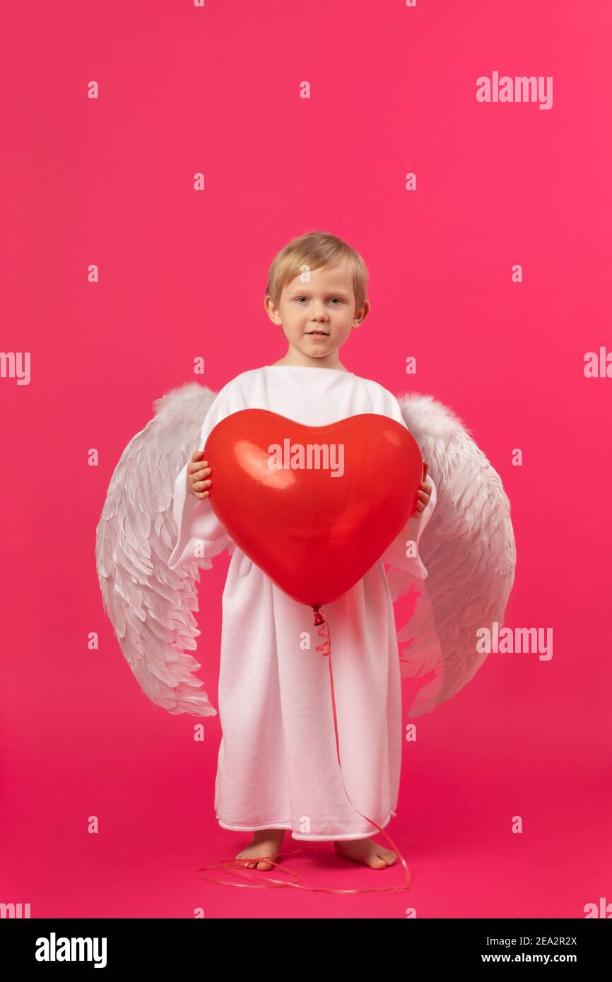Ange avec des ailes blanches tient le ballon en caoutchouc rouge en forme de coeur. Studio de photo du concept de la Saint-Valentin sur fond rose. Carte de vœux mignonne. Banque D'Images