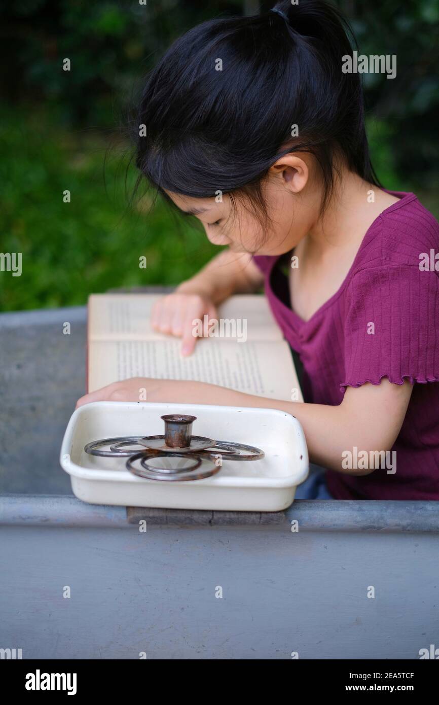 Une jeune fille asiatique mignonne assise dans une baignoire en métal antique dans un jardin lisant un livre tranquillement. Banque D'Images