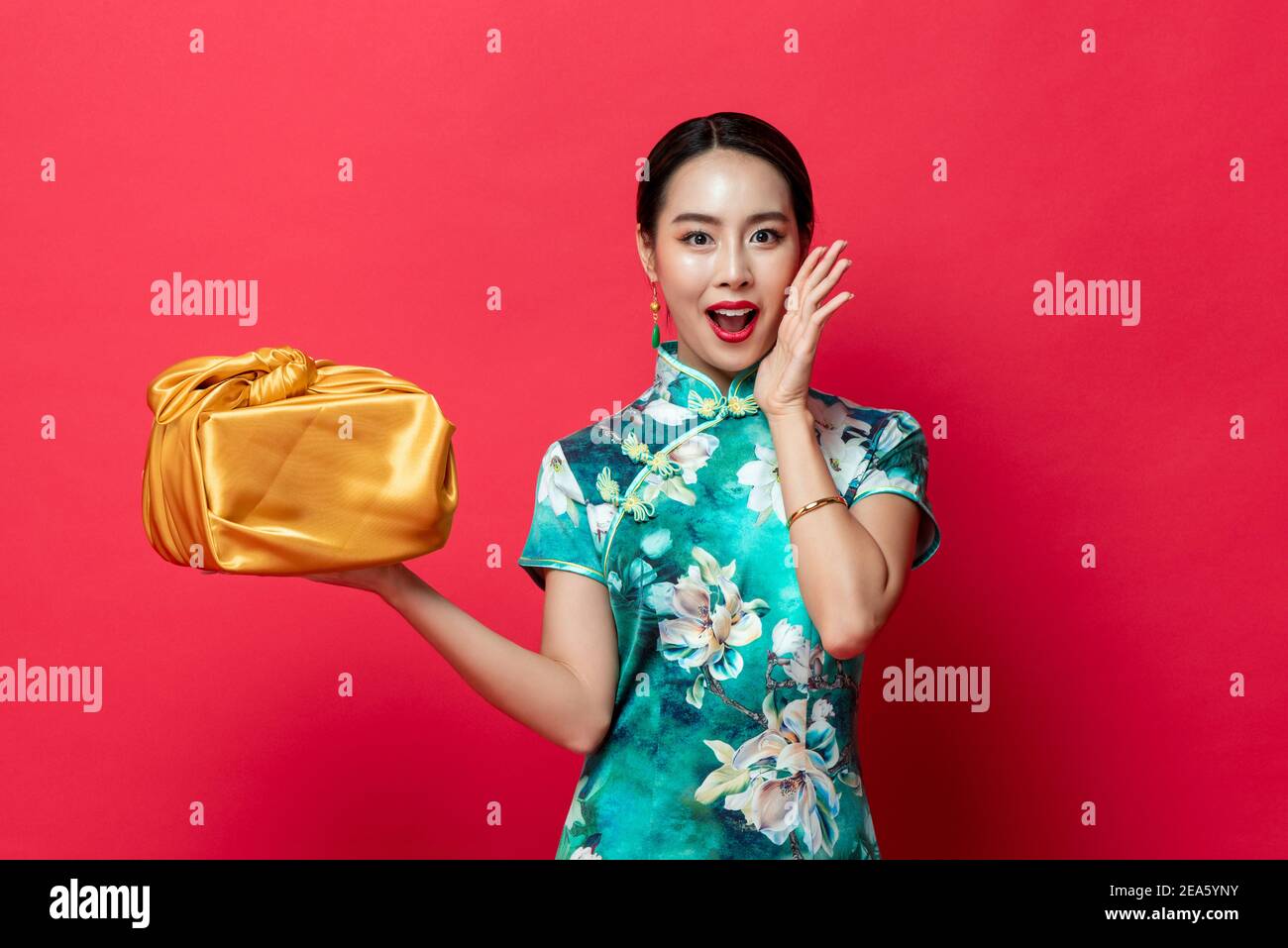 Surprise asiatique femme dans vert traditionnel Cheongsam robe tenant l'or Boîte cadeau dans un studio isolé arrière-plan rouge pour Lunar nouveau concepts de l'année Banque D'Images