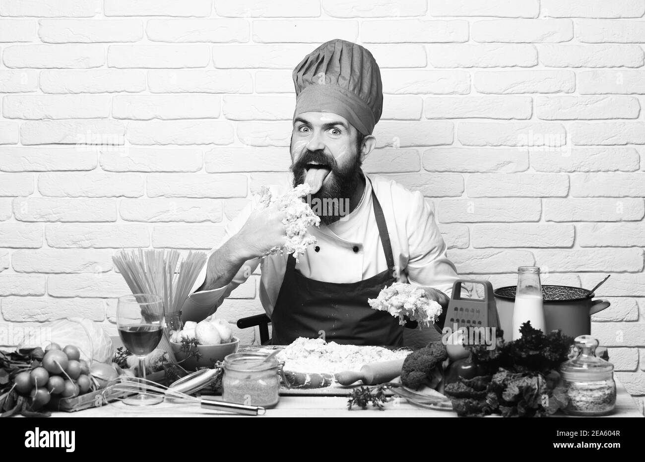 Le chef pétrit la pâte. Faites cuire avec un visage excités en uniforme bordeaux, assis près d'une table avec des légumes et des ustensiles de cuisine. Concept du processus de cuisson. Homme avec la barbe léchant les doigts de pâte sur fond de brique blanche Banque D'Images