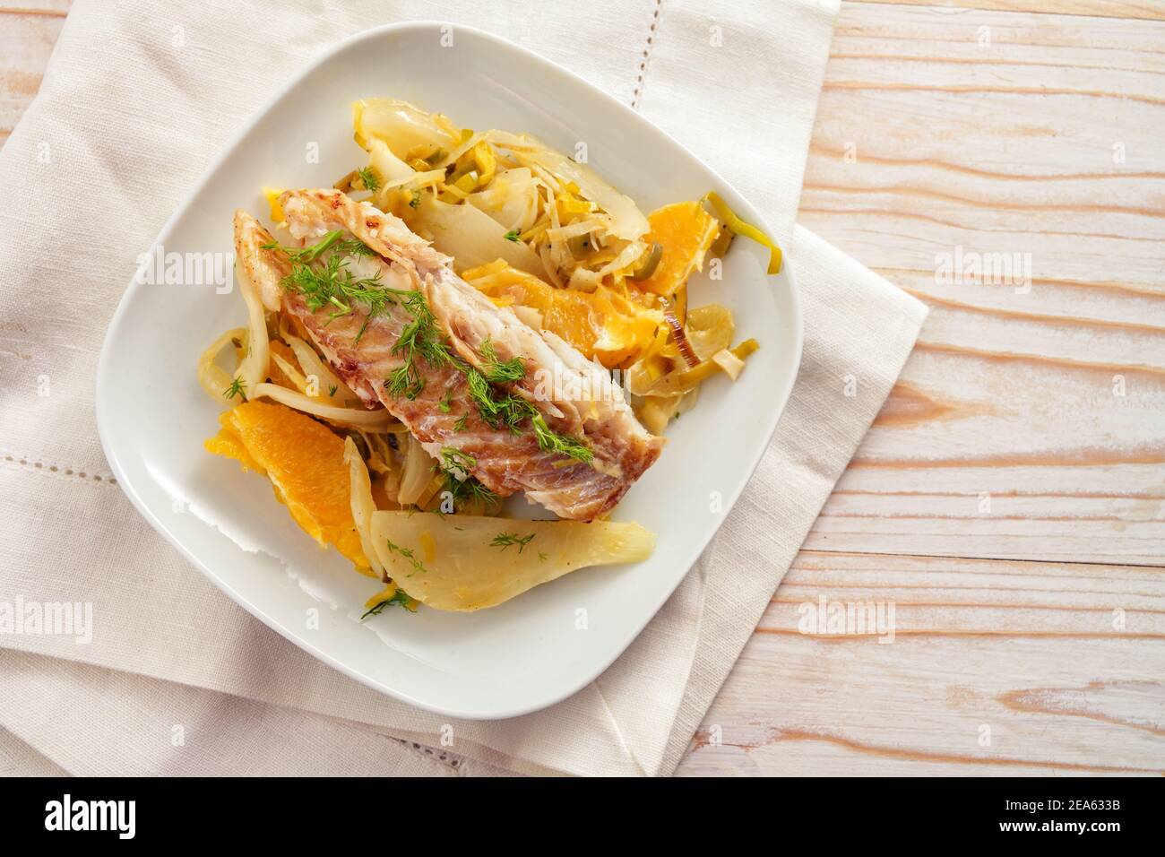 Filet de poisson de morue sur légume de fenouil avec tranches d'orange et garniture verte de fenouil sur une assiette blanche et une serviette, table en bois clair avec espace de copie, haut Banque D'Images