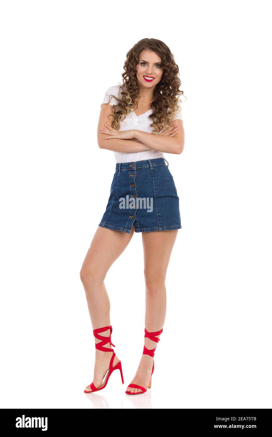 Belle jeune femme souriante en Jean mini jupe, blanc haut et rouge talons hauts est debout avec les bras croisés. Vue avant. Prise de vue en studio pleine longueur iso Banque D'Images