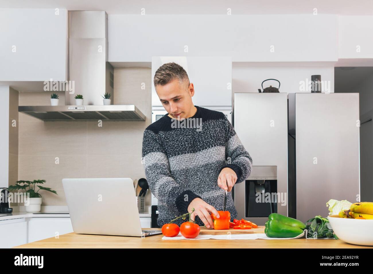 Jeune homme cuisant et préparant un repas sain dans sa cuisine et regardant des recettes vidéo sur le bloc-notes. Préparer des aliments sains, couper des légumes Banque D'Images