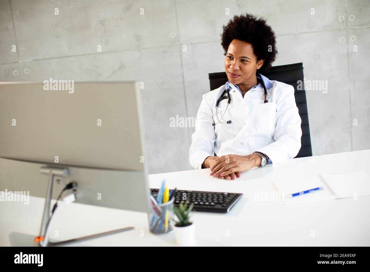 Femme afro-américaine médecin portant un manteau blanc avec stéthoscope assis derrière le bureau Banque D'Images