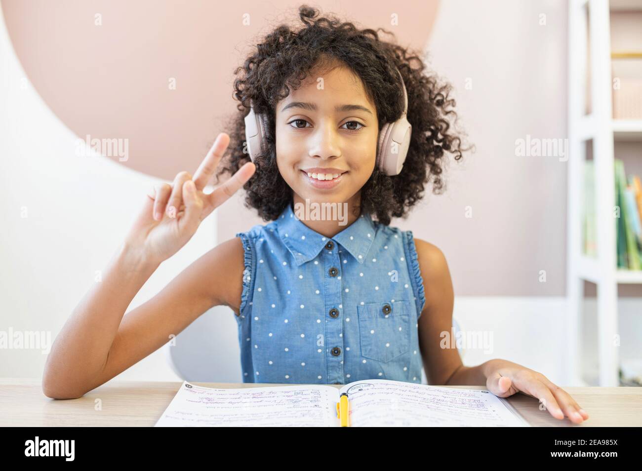 Chat vidéo avec une jeune fille africaine adorable, vêtue d'un casque, elle regarde dans l'appareil photo, montre le geste de la victoire et sourit. Un joli biracial Banque D'Images